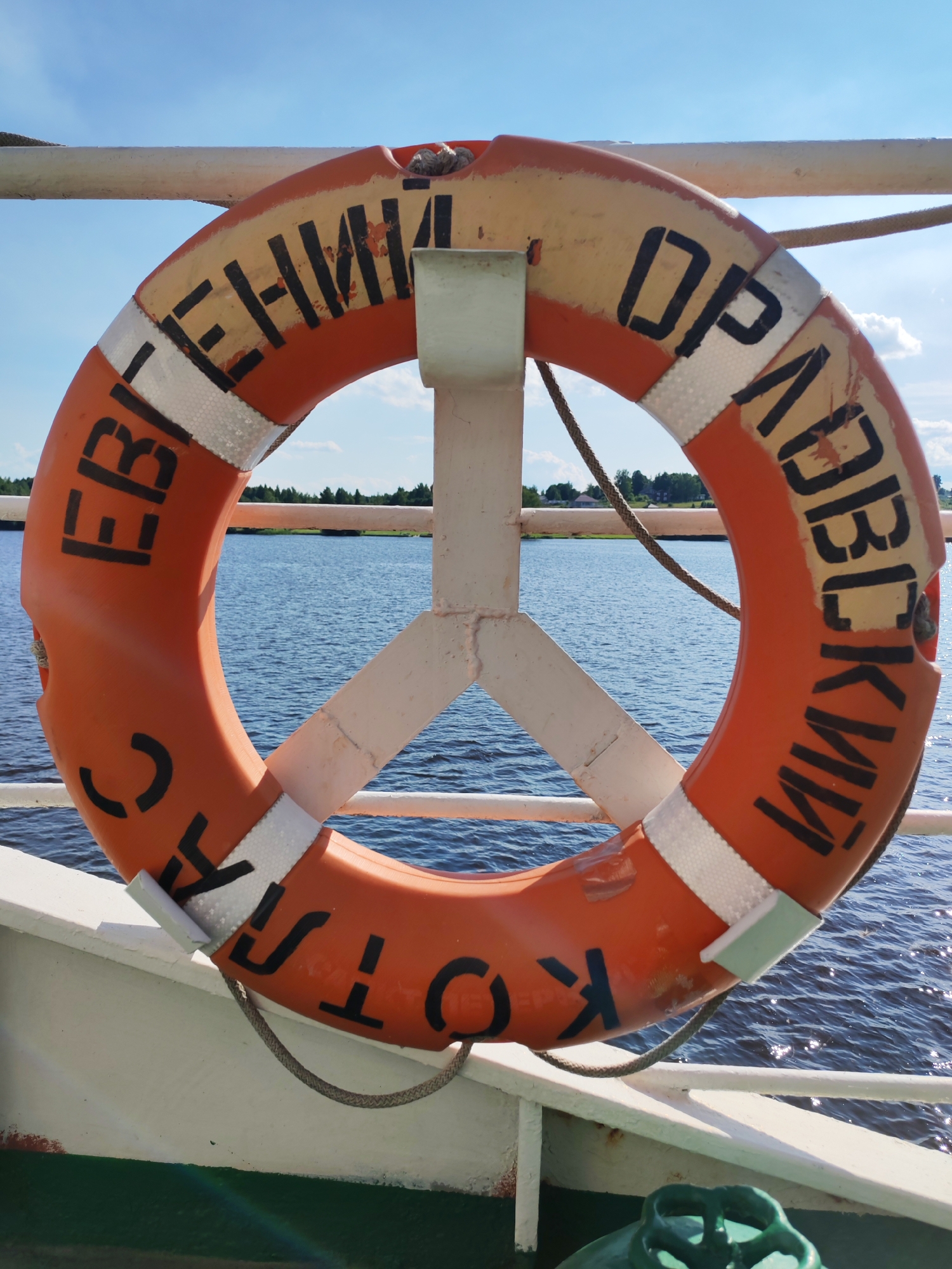 The image shows an orange life preserver hanging on a railing of a boat. The life preserver has black lettering on it that appears to be Russian. The letters "Евгений" are visible on the left side, and "Дравоскии" is visible on the right side. The bottom of the life preserver shows "Кота" and what looks like "огня". The life preserver is attached to a white metal support. In the background, a body of water with ripples is visible, along with a distant shoreline with trees and some houses. The sky is blue with some white clouds. The sun is shining, suggesting it is daytime. The overall scene is peaceful and suggests a calm day on the water.