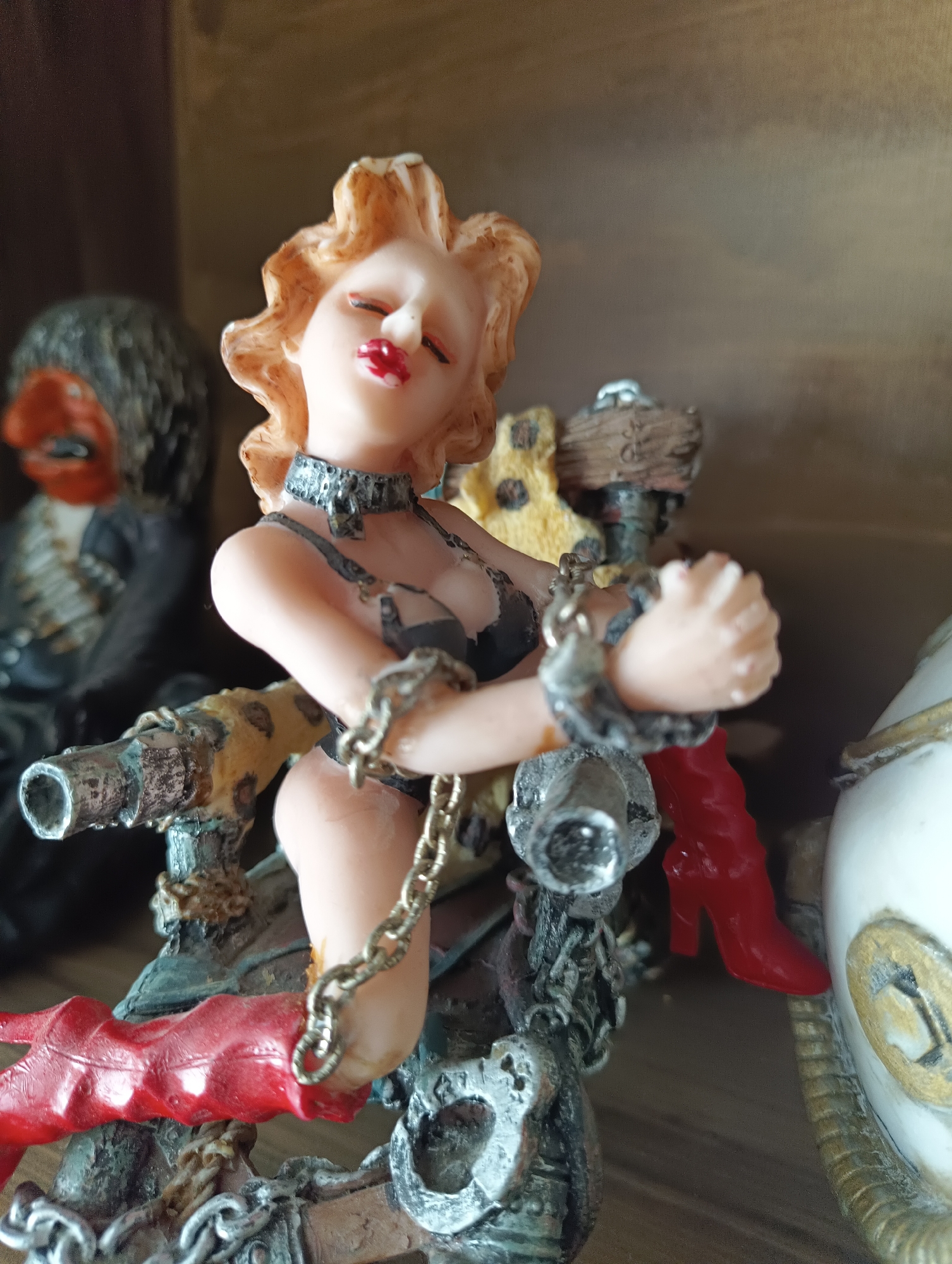 This is a close-up, low-angle shot of a detailed figurine of a woman in a provocative pose. She has blonde, wavy hair and is wearing a black choker and black lingerie. Her hands are bound by chains at the wrists and she is wearing red high-heeled boots. The figurine appears to be part of a larger, possibly elaborate, display that includes metallic and weathered elements, resembling a steampunk or gothic aesthetic. In the background, slightly out of focus, is another figurine of a person with dark hair and a red face, wearing dark clothing with decorative elements. The figurines are positioned on a shelf, indicated by the wooden background. There is no discernible information in the image that can associate this scene with the city of Karnobat, Bulgaria.