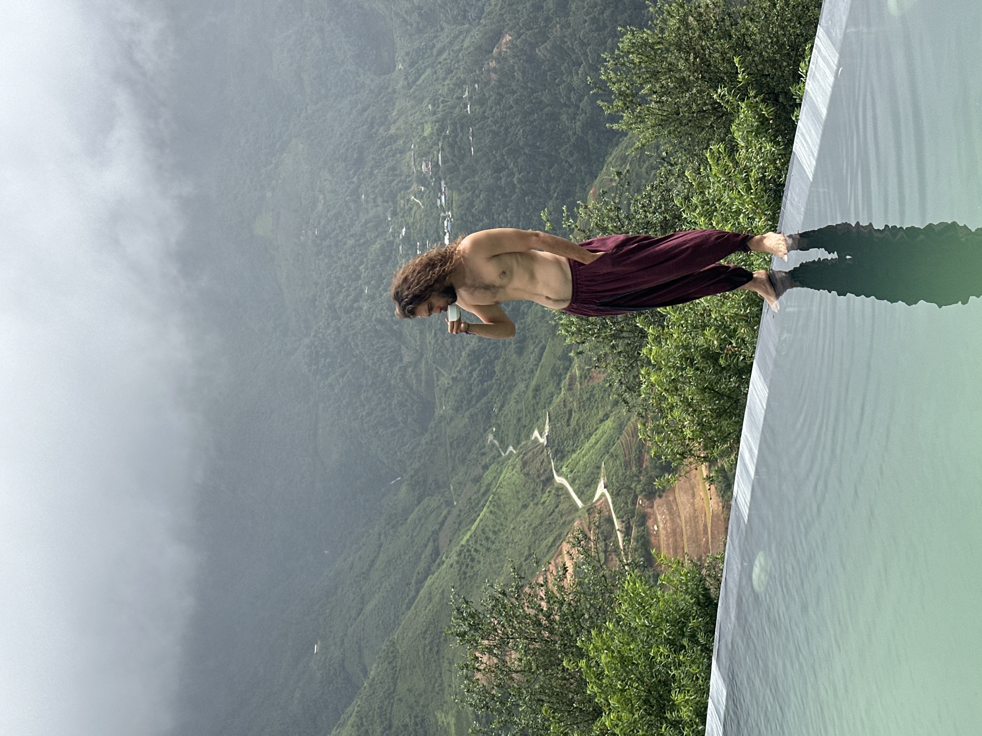 A shirtless man with curly brown hair and a beard stands on the edge of an infinity pool overlooking a misty, mountainous landscape in Ta Xua, Vietnam. He is wearing loose, maroon pants and is holding a white cup to his lips, appearing to drink. The pool's surface reflects the man and the surrounding greenery. The background features lush green mountains with a winding road visible in the distance, shrouded in fog. Trees and vegetation surround the pool and the man.