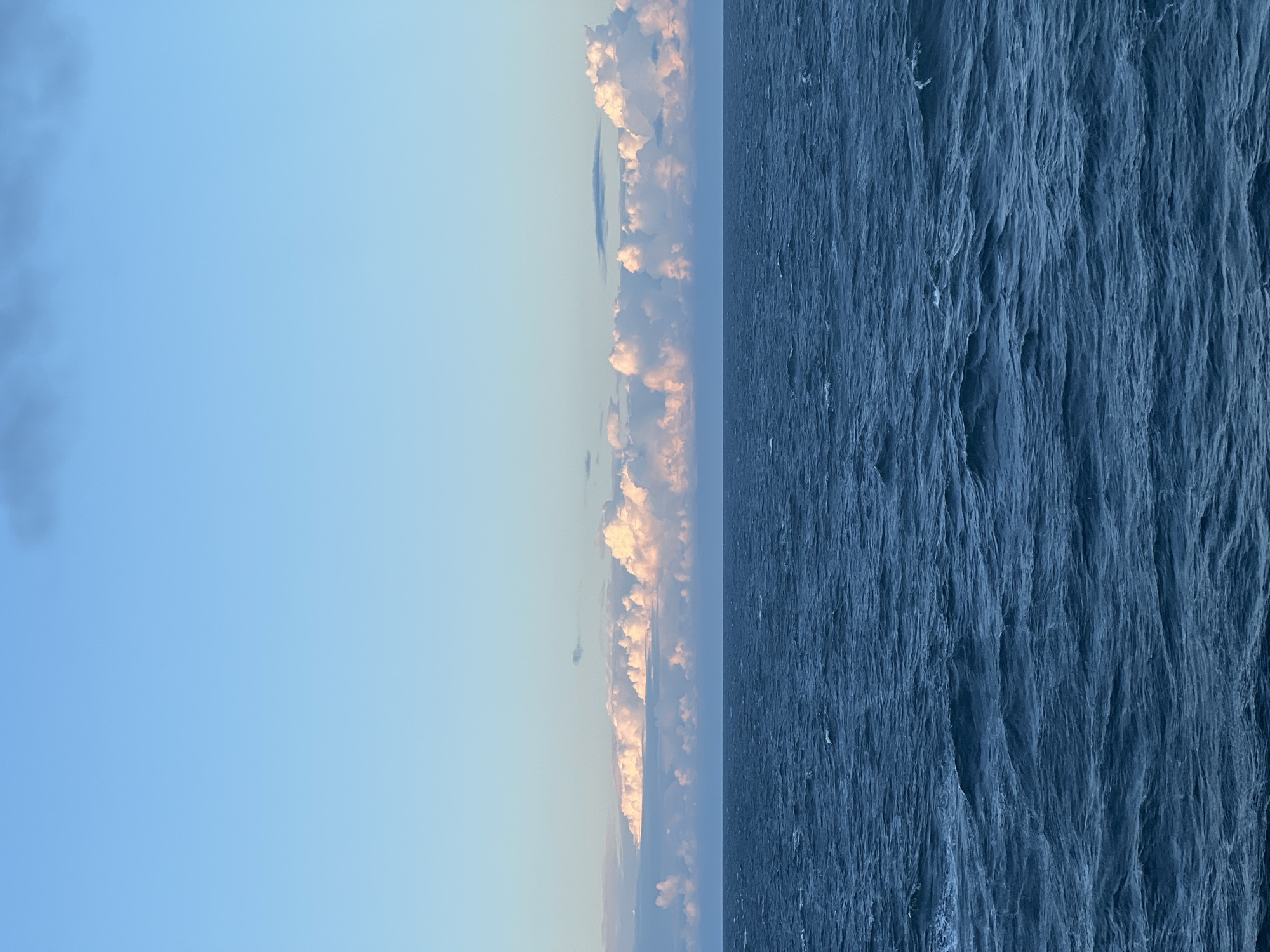 A vast expanse of dark blue, choppy ocean stretches across the lower half of the image, reflecting the sky above. The water's surface is disturbed by numerous small waves, creating a textured and dynamic appearance. A distinct horizon line divides the sea from the sky. Above the horizon, a band of fluffy white and pink clouds is illuminated by soft, warm light, suggesting either sunrise or sunset. These clouds are arranged in a horizontal line. The upper portion of the image is a clear, pale blue sky, with a few wispy, darker clouds visible towards the top edge. The overall scene is tranquil and natural, with no signs of human presence or urban elements. The location is described as Unknown, Poland, but the visual evidence suggests an open ocean environment, possibly far from any land.