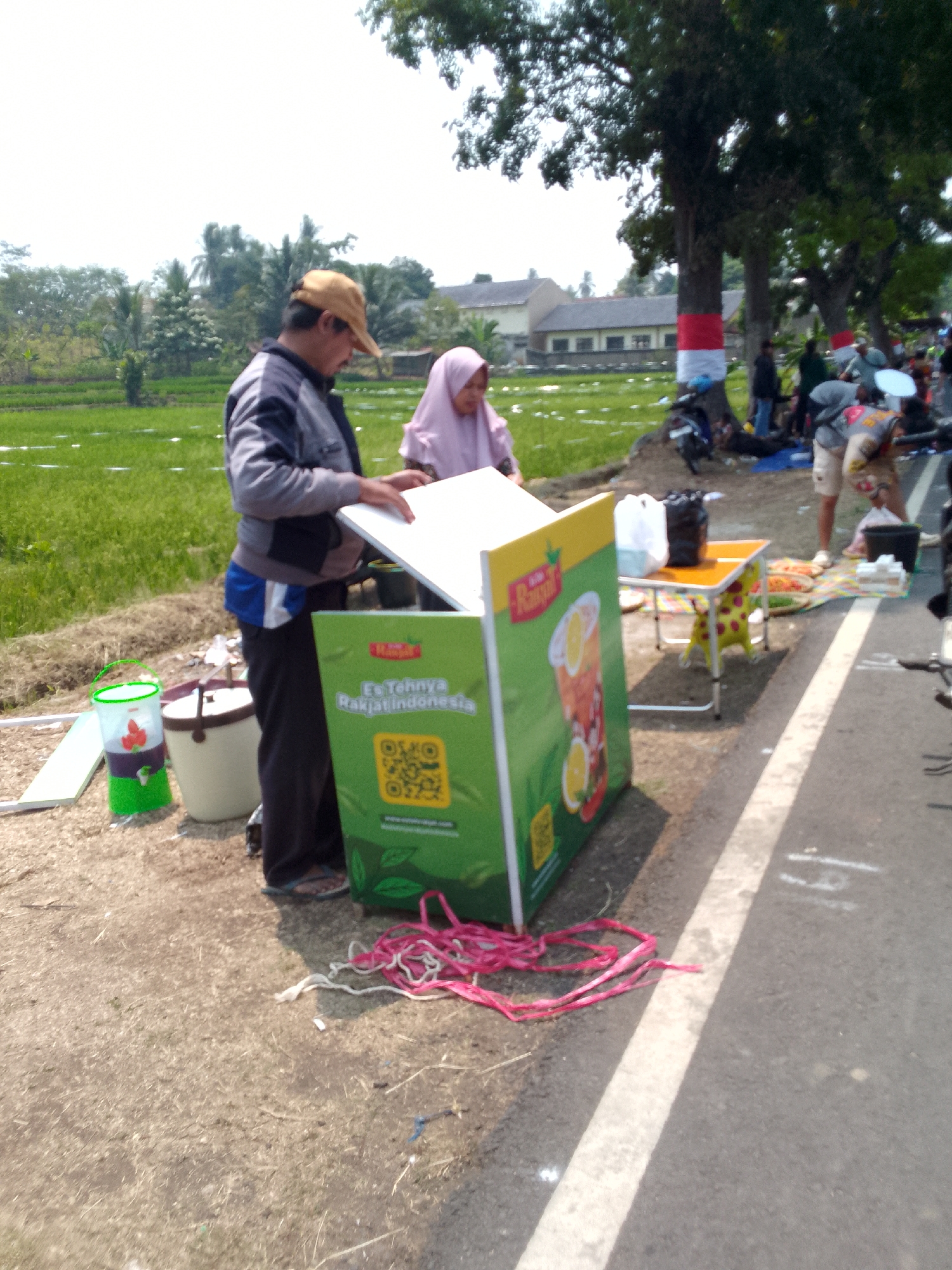 In Mandalawangi, Indonesia, an outdoor scene unfolds under a bright sky with scattered trees and green fields. A man wearing a tan cap and a grey jacket with blue trim stands to the left, facing a large green display board advertising "Es Tehnya Rakyat Indonesia." He is holding a white board or panel, possibly a prop for the display. To his right, a woman in a pink hijab is partially visible. In front of the display, a green drink dispenser with strawberries on it sits next to a cream-colored cooler with a dark lid. A pink and white rope lies coiled on the ground near the display. Further to the right, a small orange table with red strawberry designs on its legs is set up, holding two bags, one black and one white. Beyond the table, a colorful mat is spread on the ground with various items on it, including what appears to be food packaging and possibly drinks. A motorcycle is parked nearby. In the background, buildings and trees can be seen, with trees along the road marked with red and white striped bands. The scene suggests a roadside stall or event.