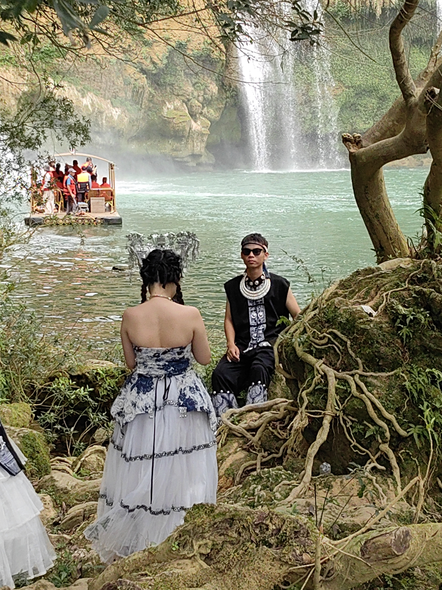 A scenic outdoor photo in Daxin County, China, features a waterfall as a backdrop to a staged scene. A boat containing approximately ten people wearing red or orange life jackets is visible on the water in the distance. To the right of the frame, a person wearing sunglasses and a black vest with decorative patterns is seated on a mossy rock formation with visible tree roots. To the left, a woman with elaborate braided hair adorned with a silver headdress stands with her back to the camera, wearing a strapless white and blue dress with multiple tiered layers. Another partially visible white dress is present on the left. The environment includes lush greenery, water, and rocky terrain, typical of a natural park setting.