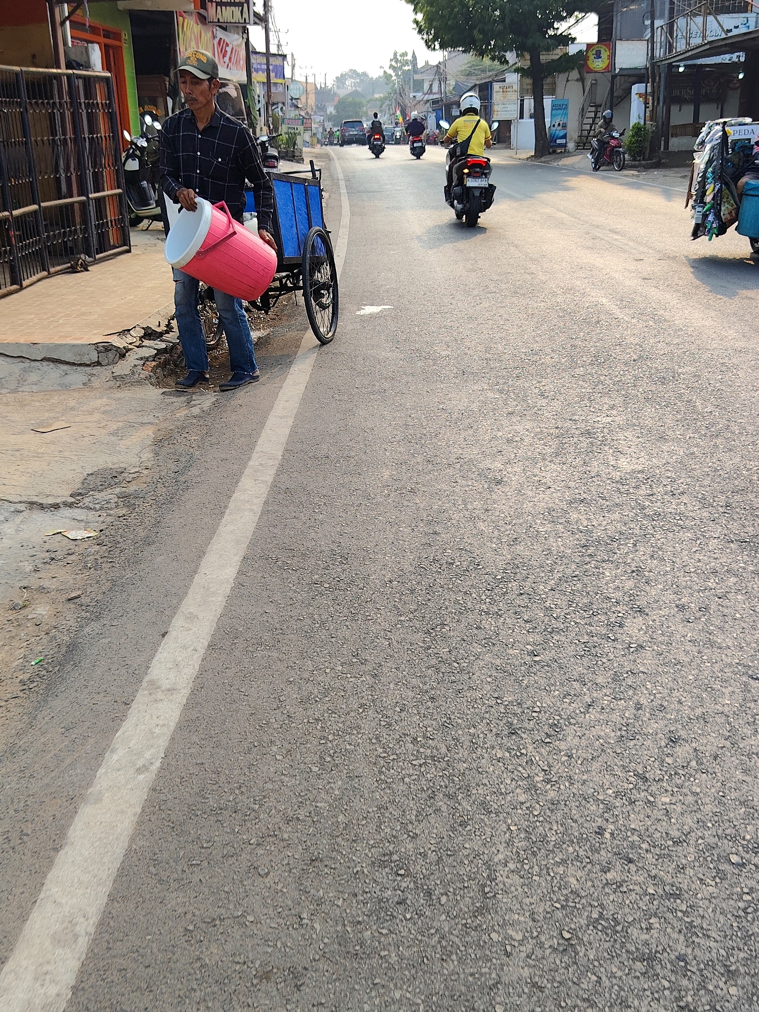 A man in a plaid shirt and jeans is standing on the side of a road in Purwakarta, Indonesia, holding a large pink insulated container. He is next to a blue cart with two wheels, one of which is visible. The man appears to be a street vendor. The road is asphalt and has a white line marking the lane. There are buildings and shops on the left side of the road, and other people on motorcycles in the distance. The sun is low in the sky, casting long shadows, suggesting it is either early morning or late afternoon. The environment appears to be an urban setting with moderate traffic.