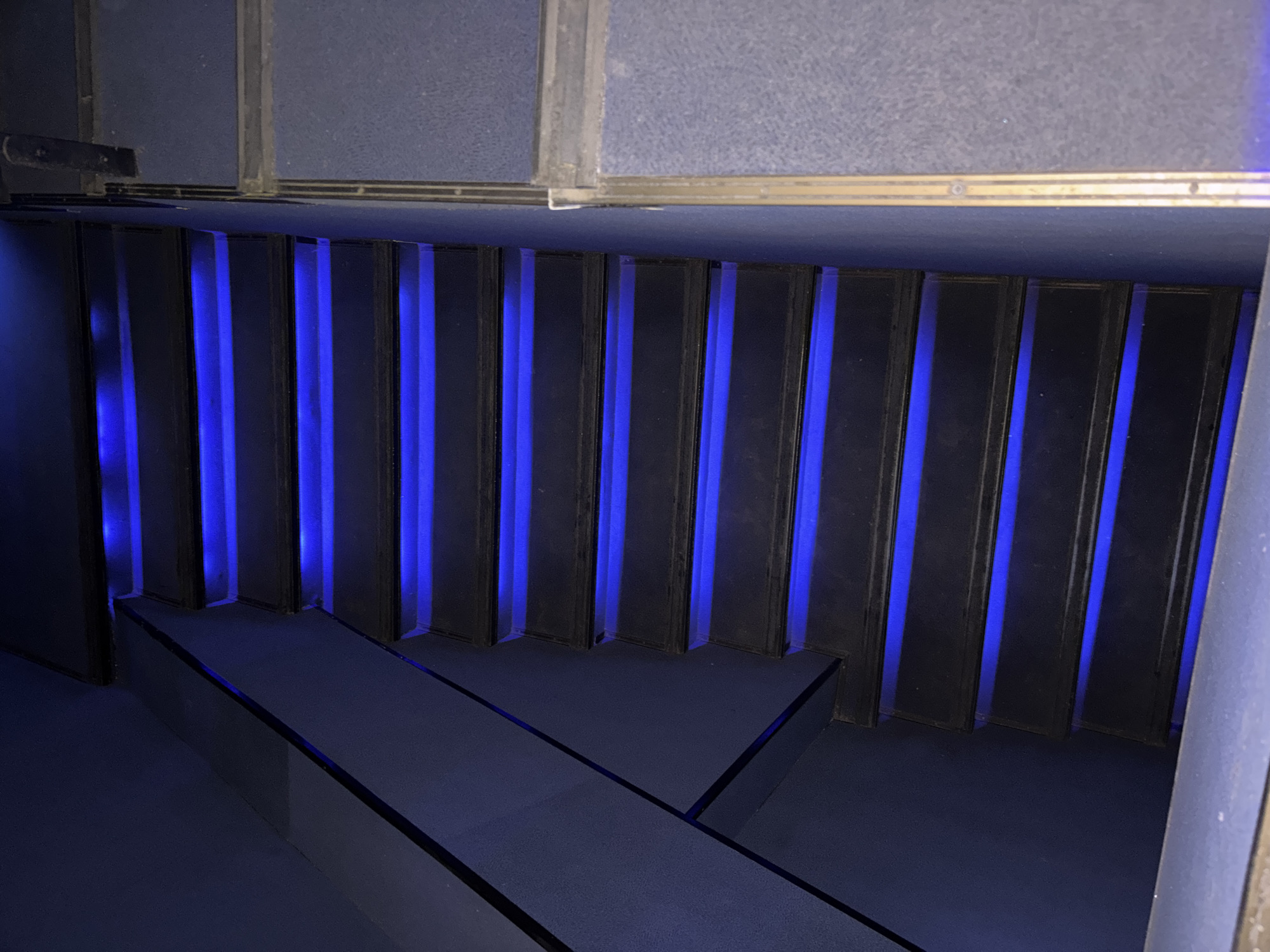 The image shows a downward view of a staircase illuminated with blue light. The steps are dark and lead down. The walls and ceiling surrounding the staircase appear to be made of gray materials. There is no indication of the location being Izhevsk, Russia or the presence of any people.