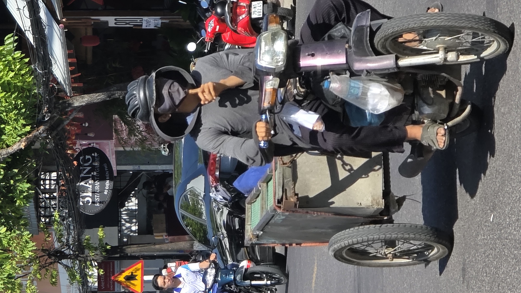 The image shows a man riding a modified motorcycle with a sidecar on a street in Bắc Nha Trang, Vietnam. The motorcycle is purple and has a headlight. The sidecar is a metal box with a wheel. The man is wearing a black helmet, a gray hoodie, black pants, and sandals. He is holding a blue bottle in his left hand. A warning road sign appears in the foreground, and a man on a scooter passes behind the modified motorcycle. In the background, there is a restaurant called "3LANG Restaurant & Garden" located at 80 Điện Biên Phủ. Other motorcycles and cars are parked or moving on the street. There are trees and power lines along the side of the road.