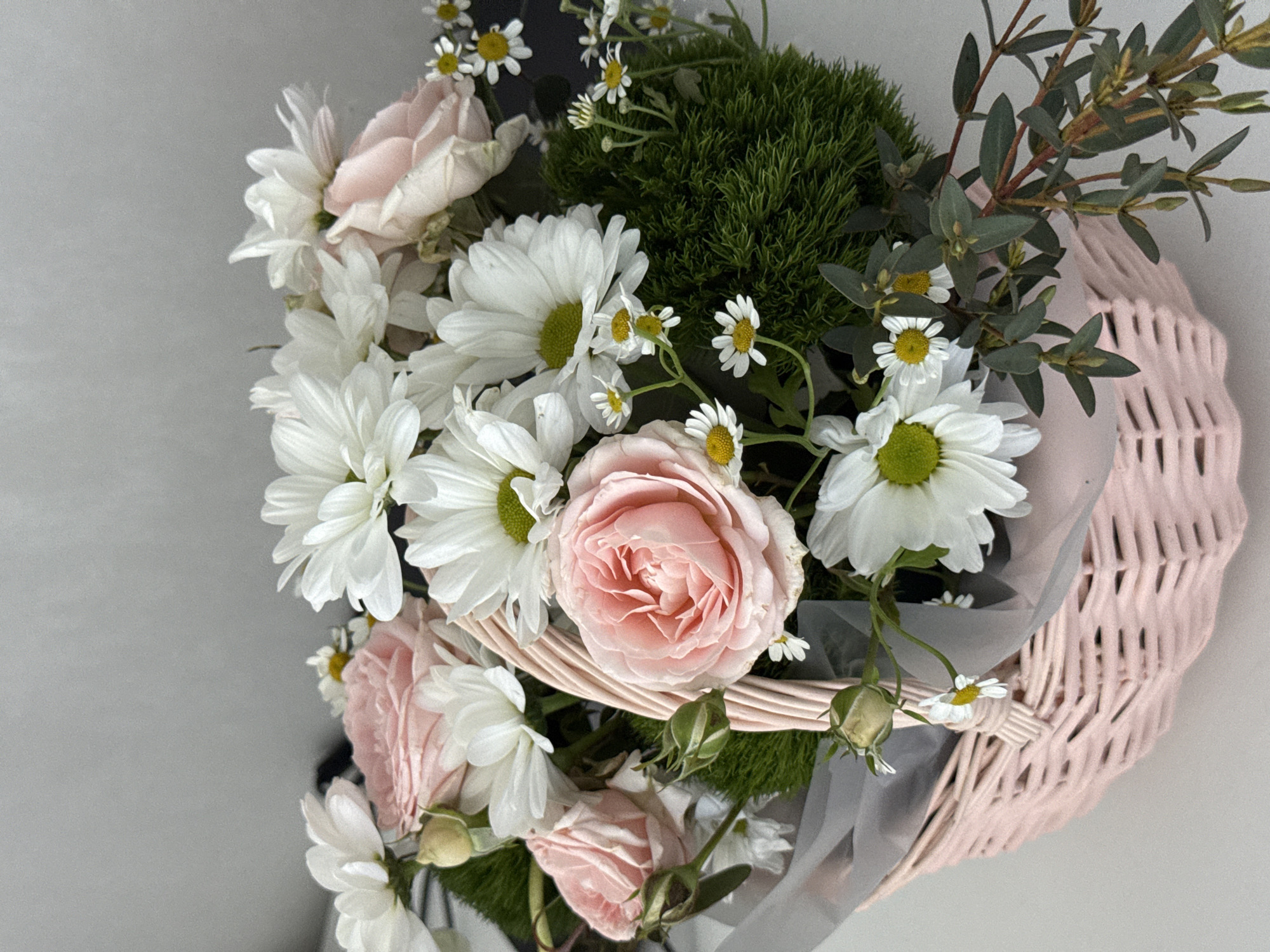 A bouquet of flowers sits in a pink wicker basket, likely on a table or flat surface. The bouquet contains a mix of white daisies with yellow centers, light pink roses in various stages of bloom, small white daisy-like flowers, and dark green foliage that appears moss-like. Some eucalyptus branches with small, rounded leaves are also present. A sheer, gray wrapping material is partially visible. The scene is well-lit. There is no information to determine that the location is in Krasnoyarsk, Russia.