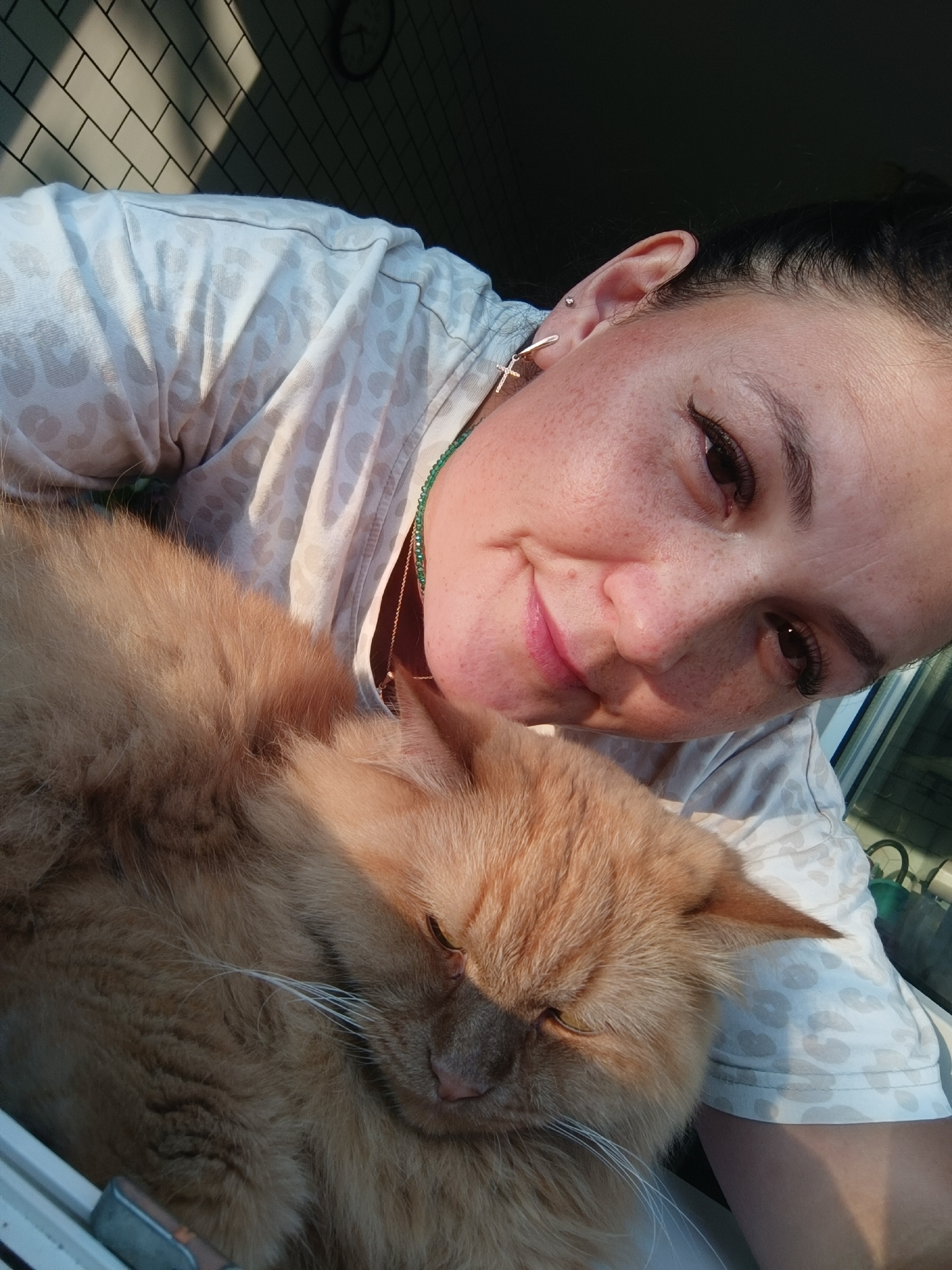 This is a close-up, self-taken photograph of a woman and her ginger cat. The woman is lying down, resting her head near the cat. She has dark hair pulled back, freckles on her face, and is wearing a light-colored shirt with a subtle leopard print. She is also wearing a cross-shaped earring and a beaded necklace. The cat is a fluffy ginger breed and appears to be sleeping or resting its head on the woman's shoulder, with its eyes closed. The background shows a tiled wall with diagonal shadows cast from sunlight, suggesting an indoor location with a window or light source. A clock face is visible on the wall in the upper left corner. There is no indication of the specific location within the city of Chaikovsky, Russia.