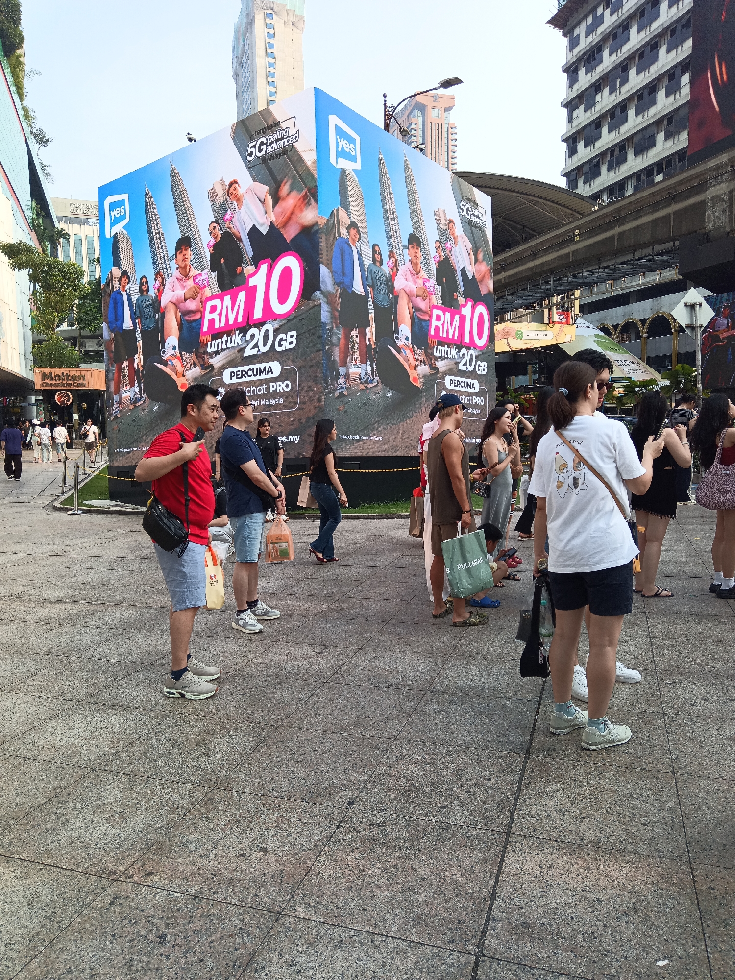The image depicts an outdoor urban scene, likely in Kuala Lumpur, Malaysia, given the prominent advertisement featuring the Petronas Twin Towers. The focal point is a large, cube-shaped advertisement displaying a promotion for "Yes" 5G, offering "RM10 untuk 20GB". Several individuals are captured standing or walking on a paved plaza.
In the foreground, a man in a red t-shirt and light blue shorts is standing, his left hand pointing towards the advertisement. To his right, another man in a dark blue t-shirt and light blue denim shorts is looking to the side. A woman in a grey dress stands further back, looking at her phone. To the right, a man in a brown tank top and shorts holds a green shopping bag.
Other individuals are visible in the middle ground and background, some engaged in conversation, others walking or looking at the advertisement. Several shopping bags and purses are carried by the people. In the background, modern buildings and an elevated train track are present, indicating a developed urban environment. The overall atmosphere appears to be a public gathering space in a commercial district.