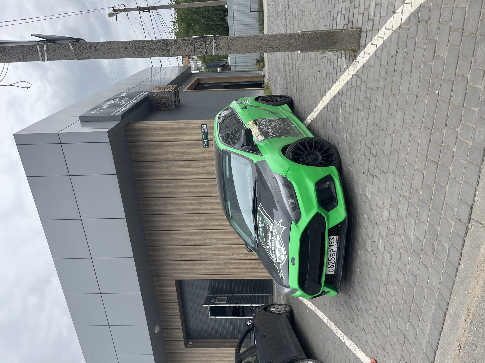 A bright green Ford Focus ST is parked on a cobblestone lot in front of a modern building. The car has a custom decal on its hood featuring a shield with checkered patterns and text. The building has a grey metal facade on the upper section and a wood-paneled lower section. A dark grey garage door is partially open, revealing a light-colored interior. A black sedan is parked to the left of the Ford Focus. A concrete pole with wires and some signage stands to the right of the car. The sky is overcast. The license plate on the Ford Focus reads "C625BP 193". The building has a sign with Russian text which appears to say "УСТАНОВОЧНЫЙ ЦЕНТР" (Installation Center). Another sign on the building reads "2046 УРАЛЬСКАЯ" (2046 Uralskaya Street). This scene is likely located on Uralskaya Street in Krasnodar, Russia.
