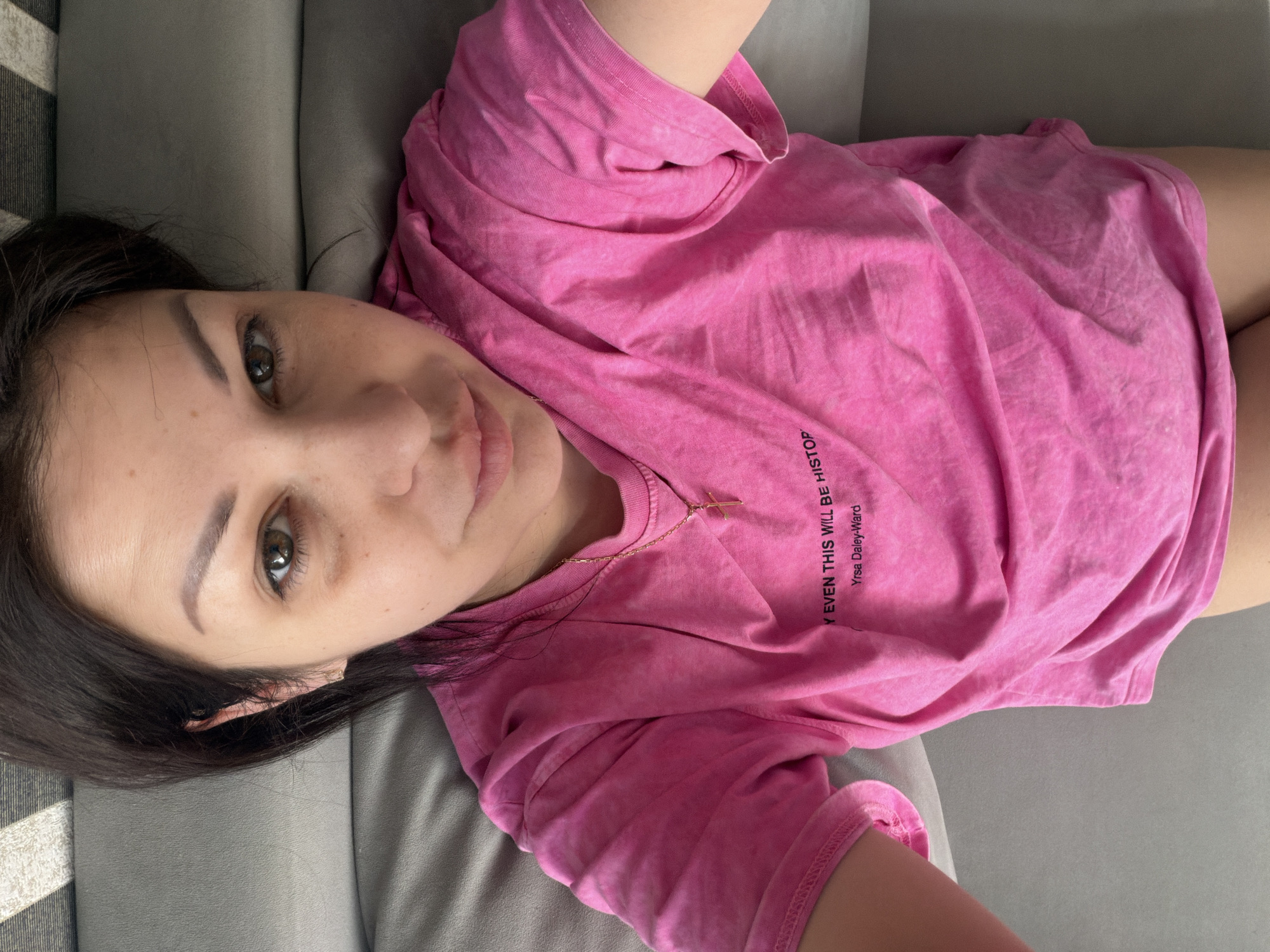 The image shows a woman lying on a grey sofa, wearing a pink t-shirt. She has dark brown hair and brown eyes. There is a gold necklace with a cross pendant around her neck. The t-shirt has text printed on it, which reads "Y EVEN THIS WILL BE HISTOR..." and "Yrsa Daley-Ward". The image is taken from a top-down perspective. There is no discernible information in the image to suggest a location in Sochi, Russia.