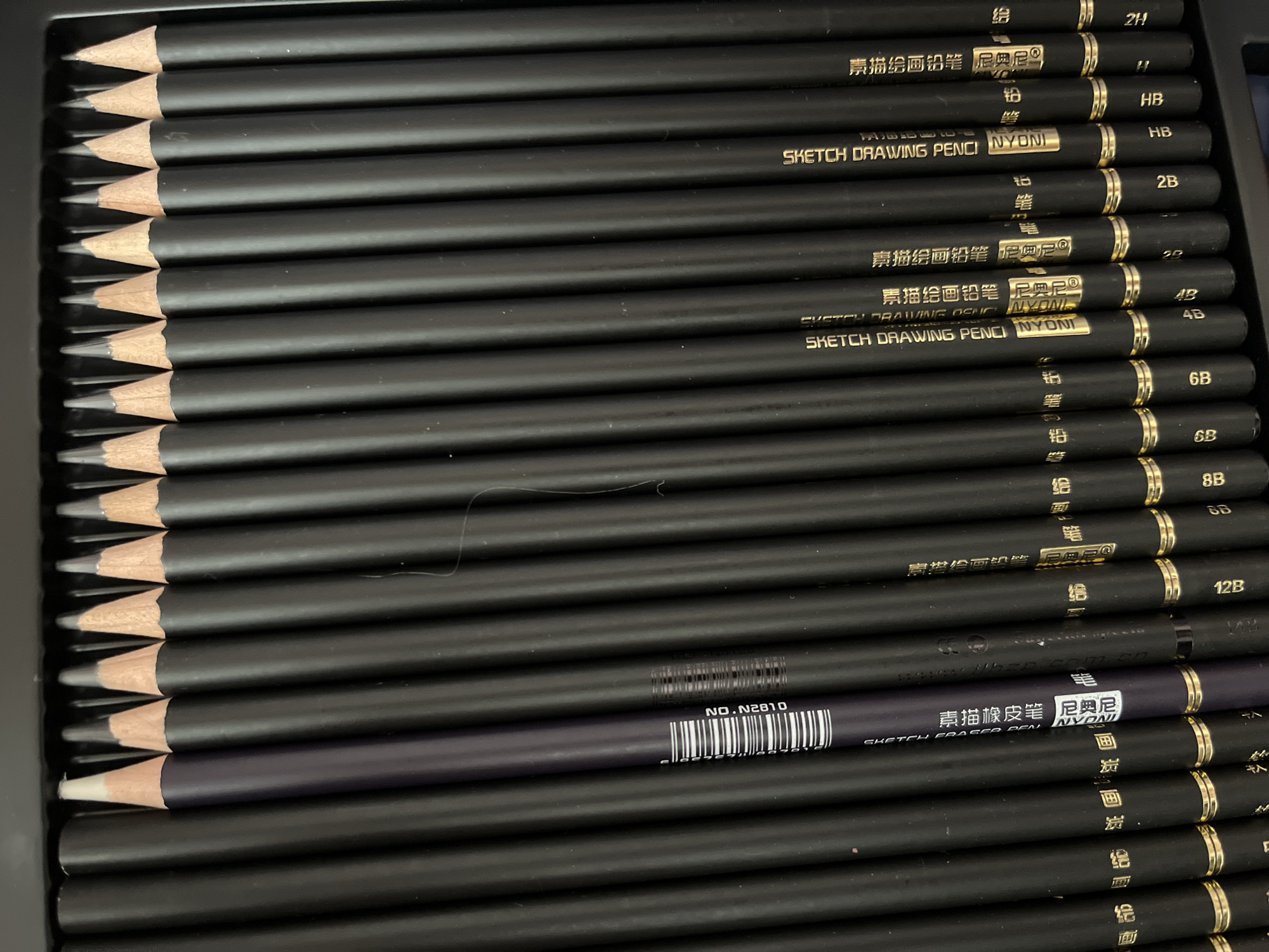 The image shows a close-up, top-down view of a collection of black drawing pencils arranged in a parallel, horizontal manner within a dark, ribbed container. The pencils have sharpened graphite tips and some have gold bands near the erasers. One pencil, slightly set apart, is a dark purple color and features a barcode and Chinese characters. Text is visible on many of the pencils, including "SKETCH DRAWING PENCIL NYONI" and grading marks like "8B", "6B", "4B", "2B", "HB", "1H", and "2H". The lighting is overhead, casting subtle shadows. There is no indication of any specific location within Krasnyye Barrikady, Russia, or any activity beyond the arrangement of the pencils.
