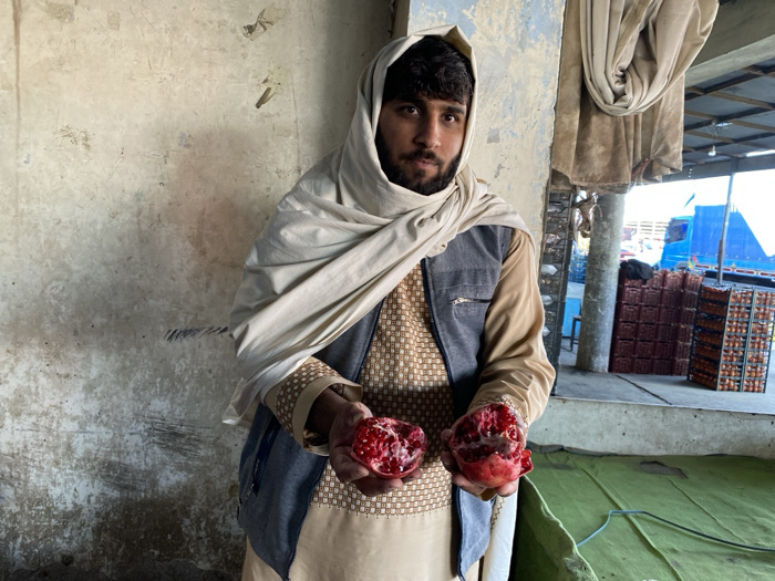 A single adult male, approximately 20-35 years old, with dark hair, beard, and mustache, stands in what appears to be a produce market or wholesale distribution center in Kandahar, Afghanistan. He is wearing a light-colored head covering, a patterned beige tunic, and a dark grey sleeveless vest. The man holds a bisected red pomegranate in each hand, presenting the fruit towards the viewer, with the arils clearly visible. He looks directly at the camera with a neutral expression. To the subject's left, a weathered, light-colored wall forms the background. To his right, an open, semi-covered area features a light blue structural column, a draped brown fabric, multiple stacks of red and brown plastic crates, and the partial view of a large blue vehicle (likely a truck) in the distance. The foreground ground on the right is partially covered by a green tarp.