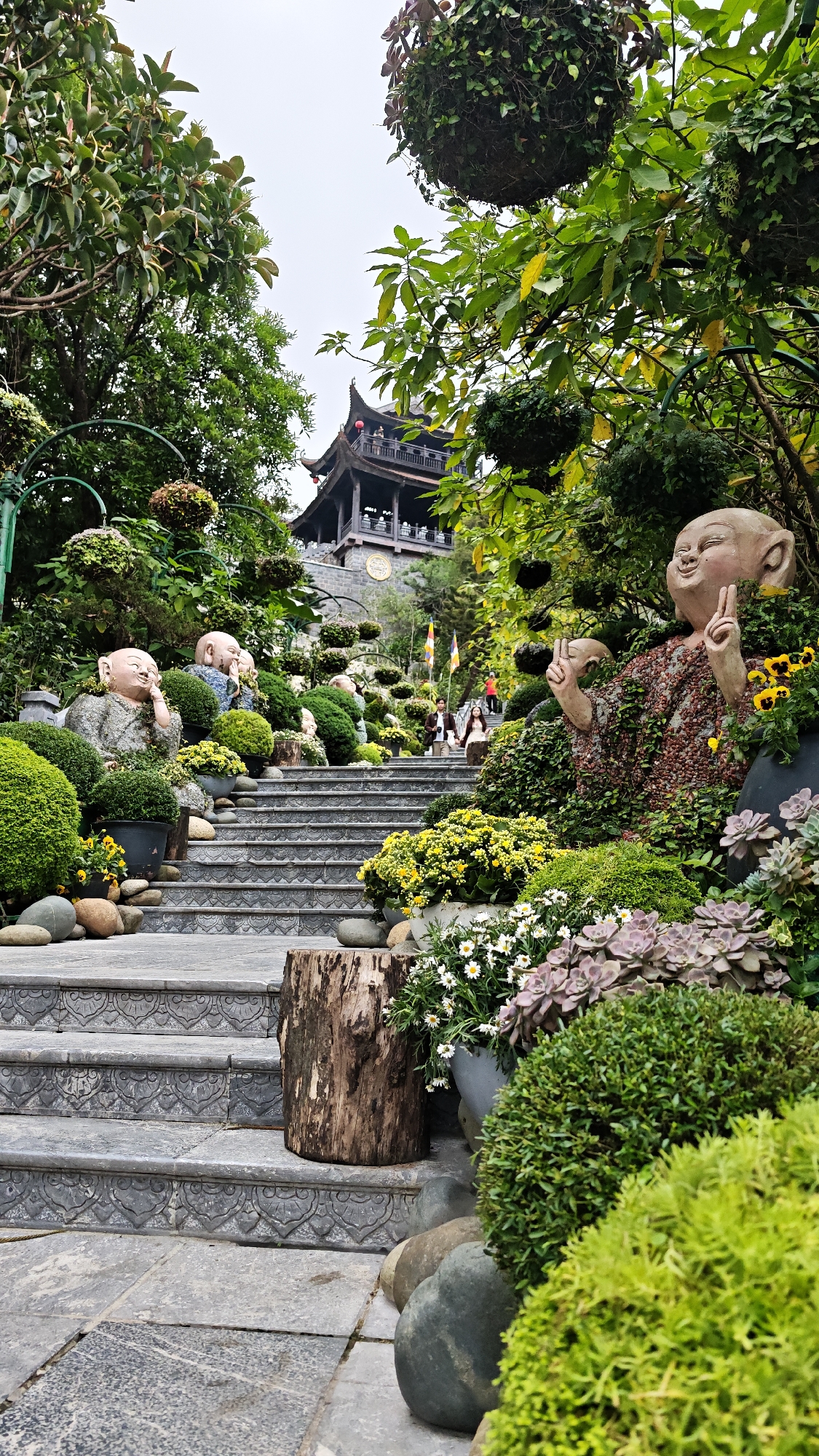 This vertical shot captures a vibrant, multi-level garden pathway ascending towards a traditional pagoda-style building, likely situated in Da Nang, Vietnam. The scene is dominated by lush greenery, colorful flowers, and a series of smiling Buddha statues.
In the foreground, large, detailed stone statues of laughing Buddhas (Hotei figures) are prominent on either side of the staircase. The statue on the right makes a "peace sign" gesture with both hands, while others along the path appear thoughtful or joyful. These statues are integrated into the landscaping, surrounded by meticulously arranged potted plants, including bright yellow kalanchoe, white daisies, and various succulents and compact green shrubs. Decorative hanging spherical planters are suspended above.
A wide stone staircase, adorned with intricate geometric patterns on its risers, leads upward through the garden. A rustic tree stump acts as a decorative element beside the stairs in the lower center. A few people can be seen ascending the stairs in the mid-ground, indicating an active site.
At the top of the incline, a dark, multi-tiered pagoda building with traditional East Asian architectural features stands against an overcast sky. A large clock face is visible on the pagoda's lower section. Several colorful flags are also visible near the pagoda. The overall atmosphere is peaceful and serene, with a rich display of horticulture and cultural statuary. No discernible text is visible in the image.