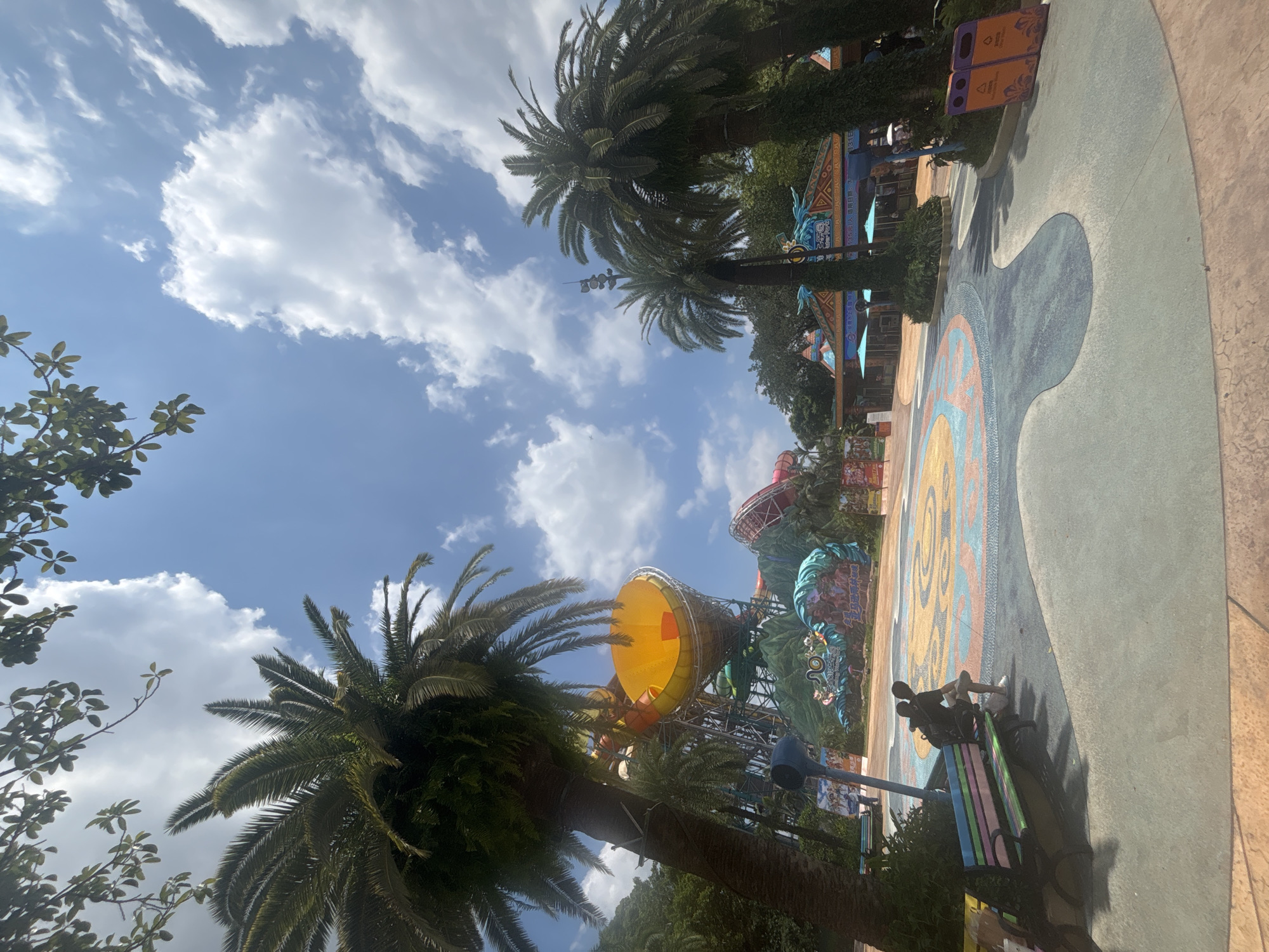 In Panyu District, China, on a sunny day with scattered clouds, this image captures a scene at a water park. Dominating the foreground are several tall palm trees, their fronds reaching towards the blue sky. To the left, a large, bright yellow water slide funnel is visible, attached to a complex structure of metal supports and other colorful water slides.
In the middle ground, a vibrant, patterned pavement area features a colorful design with wavy blue and orange lines. On the left side of this area, a person is seated on a multi-colored park bench, facing away from the camera. Behind the bench, dense green foliage is present.
Further back, an entrance to the water park is visible, featuring a structure with colorful, patterned awnings. Two blue umbrellas are set up in front of the entrance. Two orange trash receptacles are positioned on the right side of the image, near the park's paved pathway. The overall environment suggests a recreational area within a tropical or sub-tropical climate, likely during daylight hours.