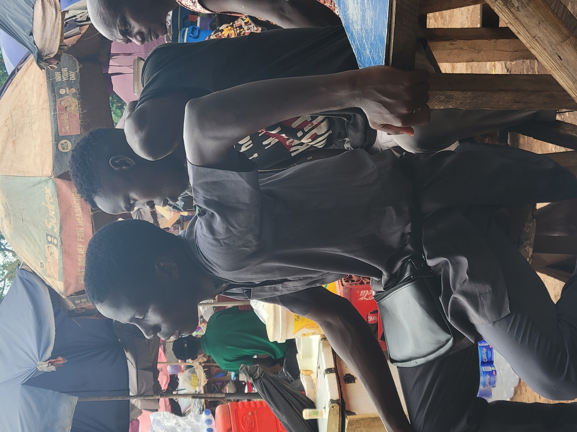 The image shows three men in a possible marketplace setting in Abuja, Nigeria. The foreground features a man in dark clothing, including pants and a buttoned shirt. He has a dark-colored bag slung across his body. He's positioned near a table, possibly a vendor's stall, where items like bottles of water are visible. Behind him and to the left is another man wearing a green shirt. Above and to the left, another man wearing black looks downward. The environment is a bustling market with stalls covered by umbrellas with faded advertisements. The background is filled with various goods, containers, and other market items, suggesting a lively commercial atmosphere. The image appears to have been taken in daylight.