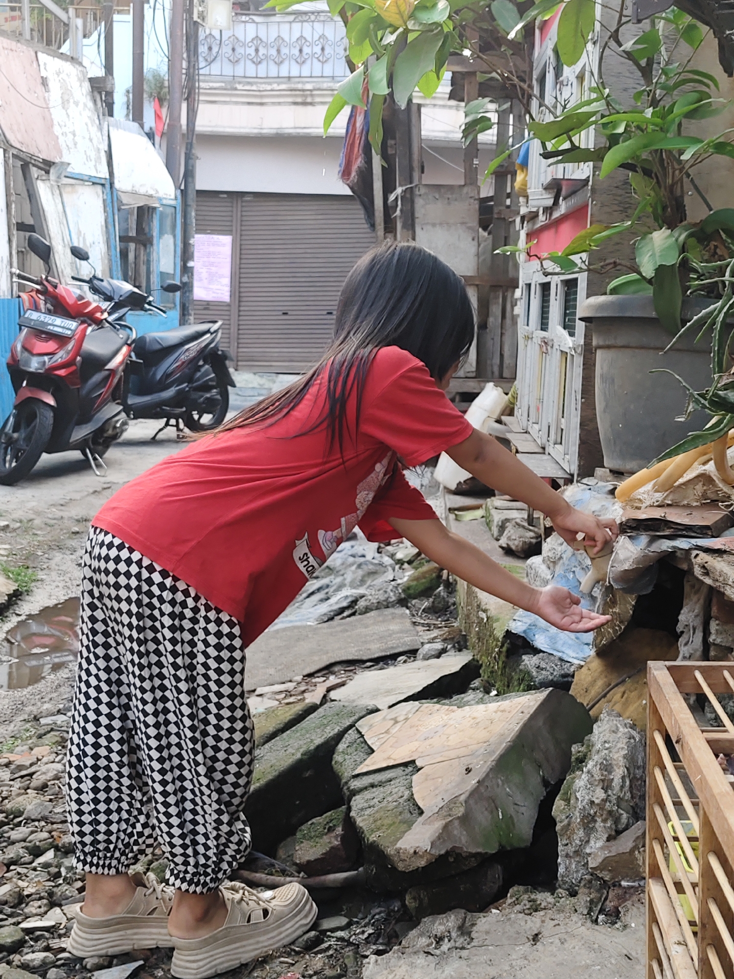 A young Indonesian girl, wearing a red t-shirt and black and white checkered pants, is bending over to reach for something. She is standing on a rocky, debris-strewn path in an urban alleyway. Behind her, a red and black motorcycle is parked. The environment suggests a somewhat neglected area with peeling paint and exposed construction materials on the buildings. The girl's hands are extended towards a small stream of water flowing over rocks, indicating she might be playing with the water or trying to catch something. The scene appears to be in South Jakarta, Indonesia, based on the presence of what looks like typical Indonesian urban architecture and the style of the motorcycle.