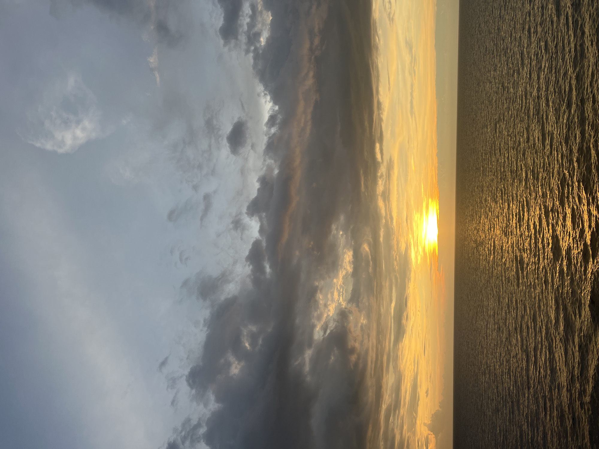 The image shows a sunset over a body of water. The sun is low on the horizon, casting a warm golden glow across the scene. The sky is filled with dramatic clouds, ranging in color from dark gray to light orange and yellow. The water is calm, with small ripples reflecting the colors of the sunset. The horizon line is sharp and distinct, dividing the sky from the water. There are no people or man-made objects visible in the image. The location is listed as Unknown, Unknown.