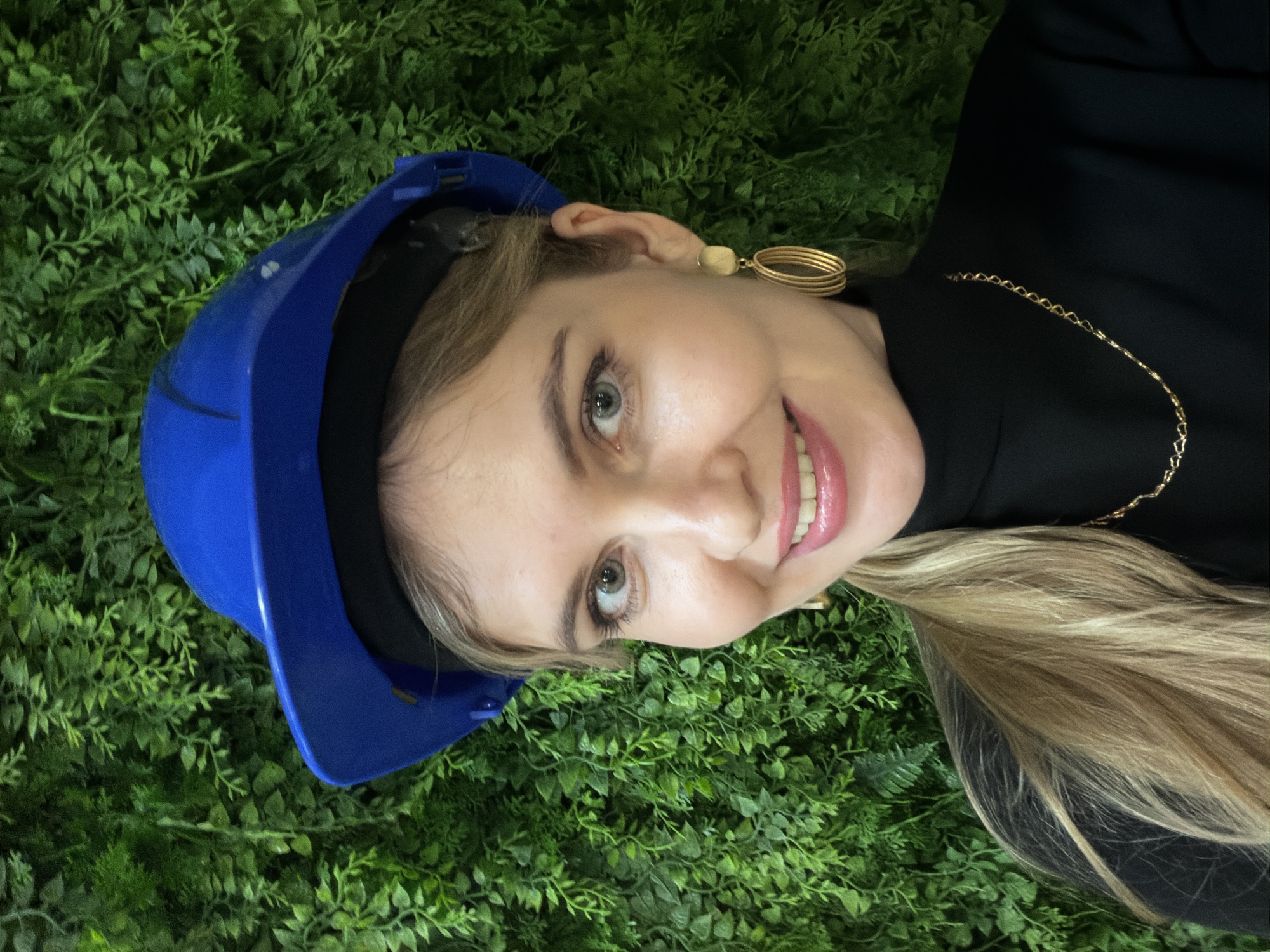 A young woman with light skin and blonde hair is wearing a blue hard hat. She is lying on a surface covered in artificial green foliage. She is wearing a black top, a gold necklace, and gold earrings. The background is a dense arrangement of small, green leaves. The location is not identifiable as Izhevsk, Russia.