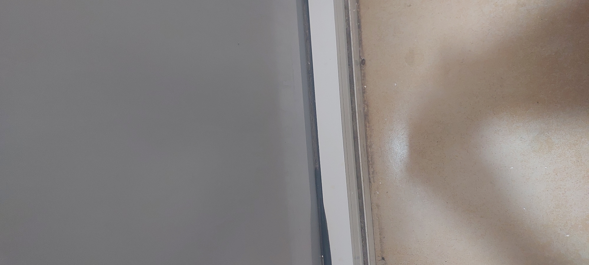The image shows a close-up of a tiled floor and the bottom edge of a wall or counter. The floor is light brown with a speckled texture. There are a few small white debris pieces scattered on the floor. Above the floor is a white horizontal strip, possibly the base of a wall or a counter. Above this white strip is a dark line, likely a sealant or shadow, followed by a plain grey surface. The scene appears to be indoors, with no indication of its specific location in Pakhtakor, Uzbekistan, beyond the general description of a floor and wall.