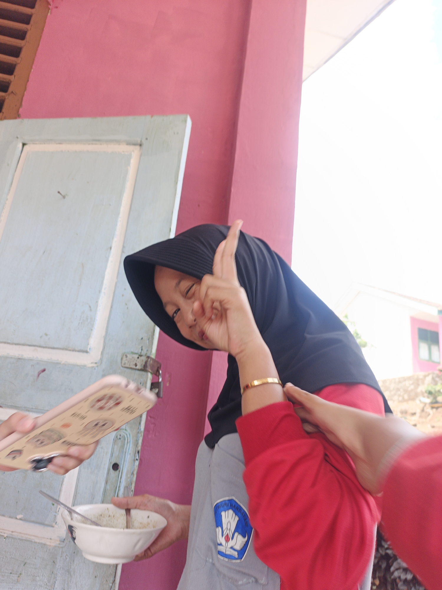 In Tanjung Setia, Indonesia, a young woman wearing a black hijab and a red long-sleeved shirt is photographed. She is holding up her right hand, forming a peace sign, and smiling at the camera. In her left hand, she is holding a white bowl with a spoon in it, which appears to contain some food. Another person's hands are visible in the foreground, holding a phone and reaching towards the woman. The background shows a pink wall and a light blue door, suggesting an outdoor setting near a building.