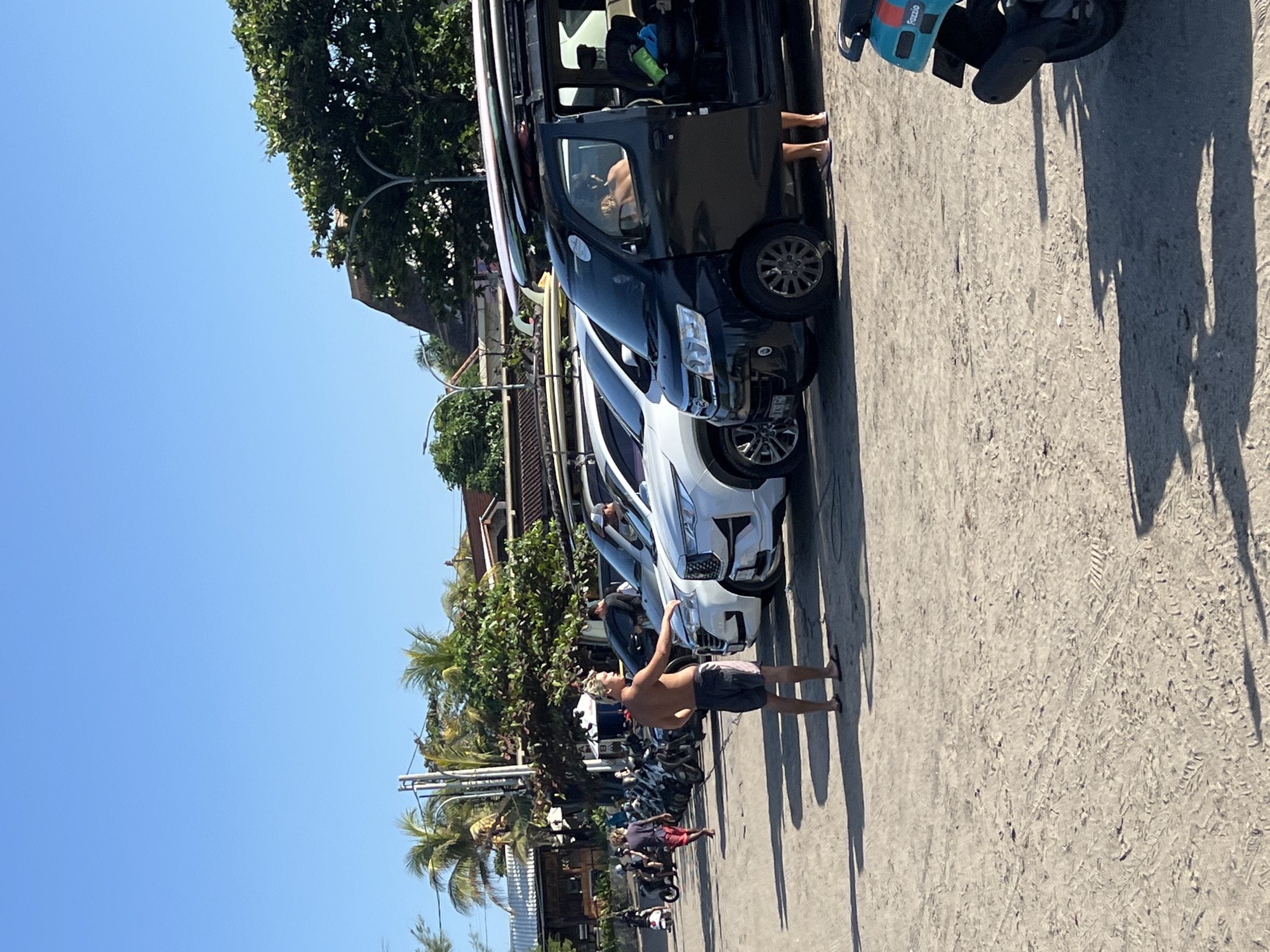 The image depicts a sunny daytime scene in North Kuta, Indonesia, likely a beach parking area. In the foreground, a shirtless man with blond hair, wearing dark shorts and flip-flops, stands facing away from the camera and towards a row of parked vehicles. He is reaching towards the hood of a white SUV. Behind him, a line of parked vehicles, including several white SUVs and a black minivan loaded with surfboards on its roof, stretches into the background. The minivan has its side door open, and a person's legs are visible inside. Further back, a line of parked scooters and motorcycles is visible, with some people walking among them. The ground is sandy, with tire tracks and footprints. Lush green trees and a few buildings with thatched roofs are visible in the background, under a clear blue sky. The overall impression is of a relaxed, possibly tourist-oriented location.