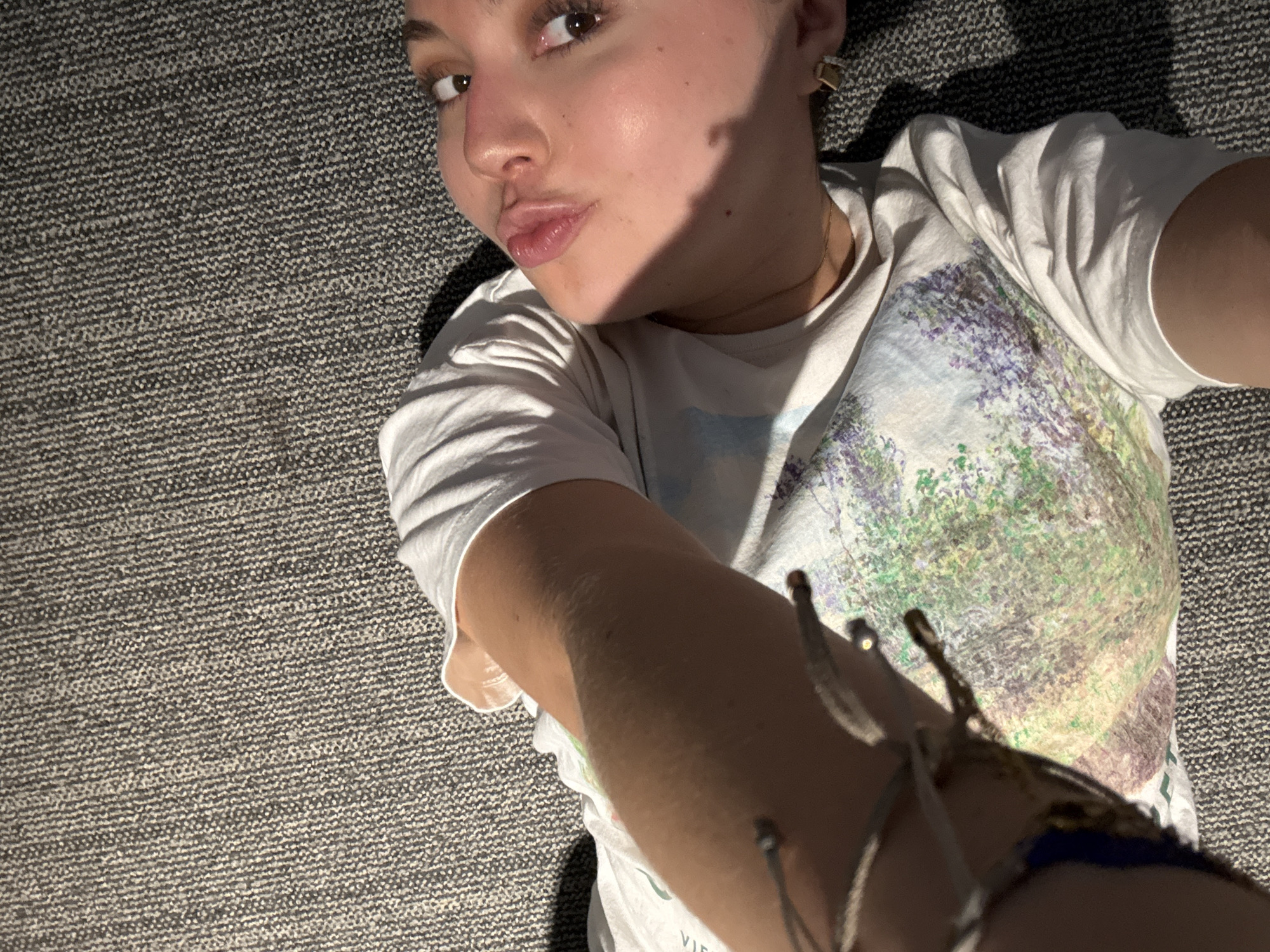 A person is lying on a gray carpet, taking a selfie. The person has light skin, brown eyes, and is wearing a white t-shirt with a floral design. A bracelet is visible on their arm. The location is unknown.
