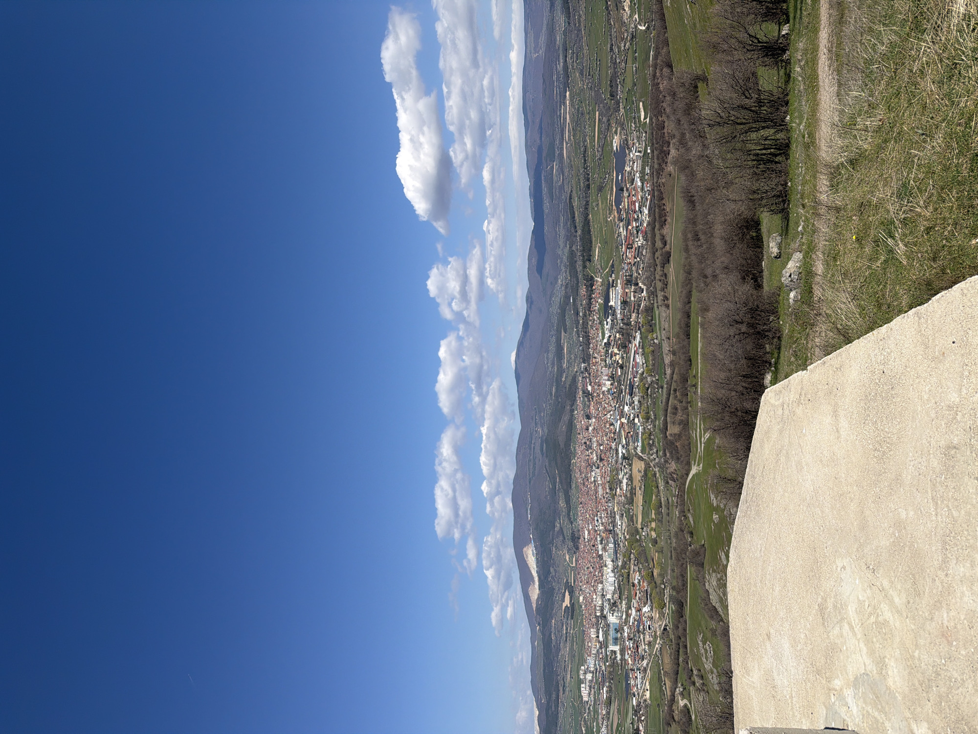The image shows a view of a city from a high vantage point. The location appears to be Bata, Bulgaria. The sky is blue with scattered white clouds. A portion of a stone or concrete structure is visible in the foreground. A town nestled between a mountain range. The slopes of the mountain are partially covered with trees and vegetation. The town has buildings with red roofs. Along the edges of the town, there are green fields. The ground is covered with grass and vegetation.