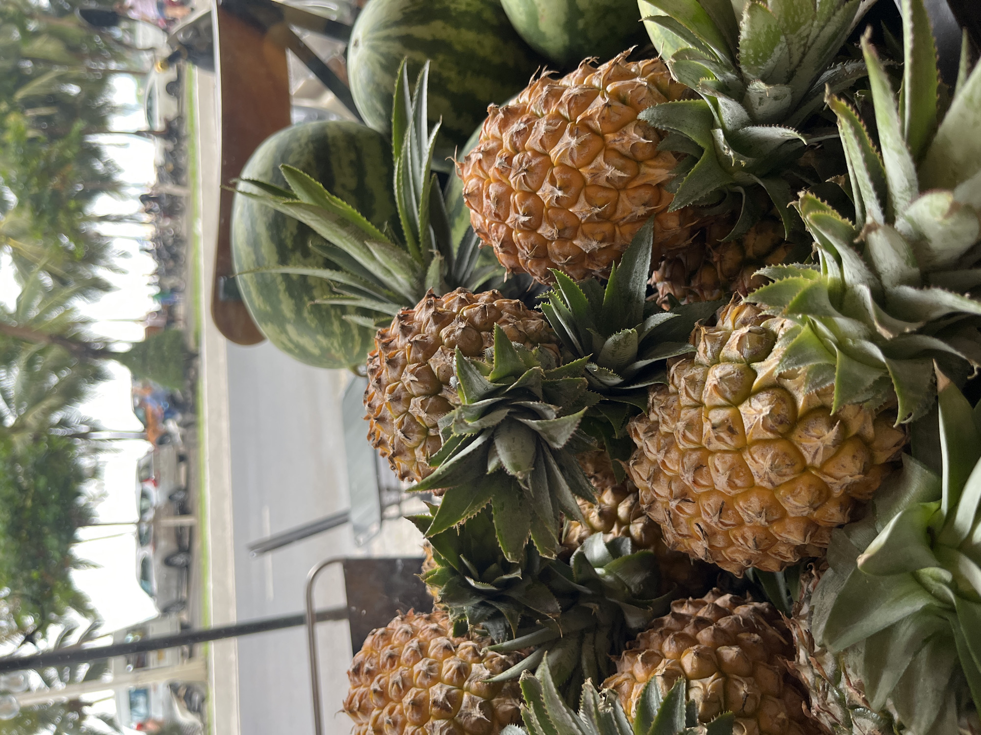 The image shows a pile of ripe pineapples and watermelons arranged on a cart in an urban environment. The pineapples are golden-brown with prickly exteriors and green, spiky tops. The watermelons are green with darker green stripes. The cart appears to be positioned near a sidewalk, with a blurry background showing trees, vehicles, and buildings, indicating a street scene. The city and specific location are not identifiable from the image.
