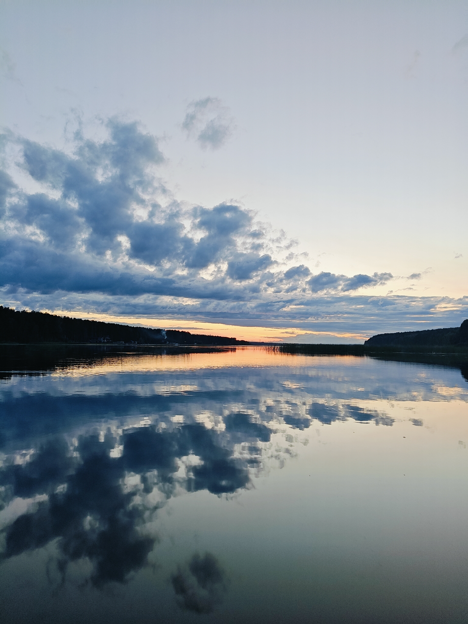 The image depicts a tranquil body of water, likely a river or lake, reflecting the sky and surrounding landscape. The time of day appears to be dusk or dawn, with soft hues of orange and yellow visible on the horizon. Dark silhouettes of trees line the far bank, creating a natural border against the fading light. The sky is partially covered by scattered clouds, which are also mirrored in the still water, creating a symmetrical effect. The overall impression is one of peace and natural beauty. While no specific landmarks are identifiable in this particular view, such serene waterfront scenes are common in and around Chaykovsky, Russia, a city situated on the Kama River.