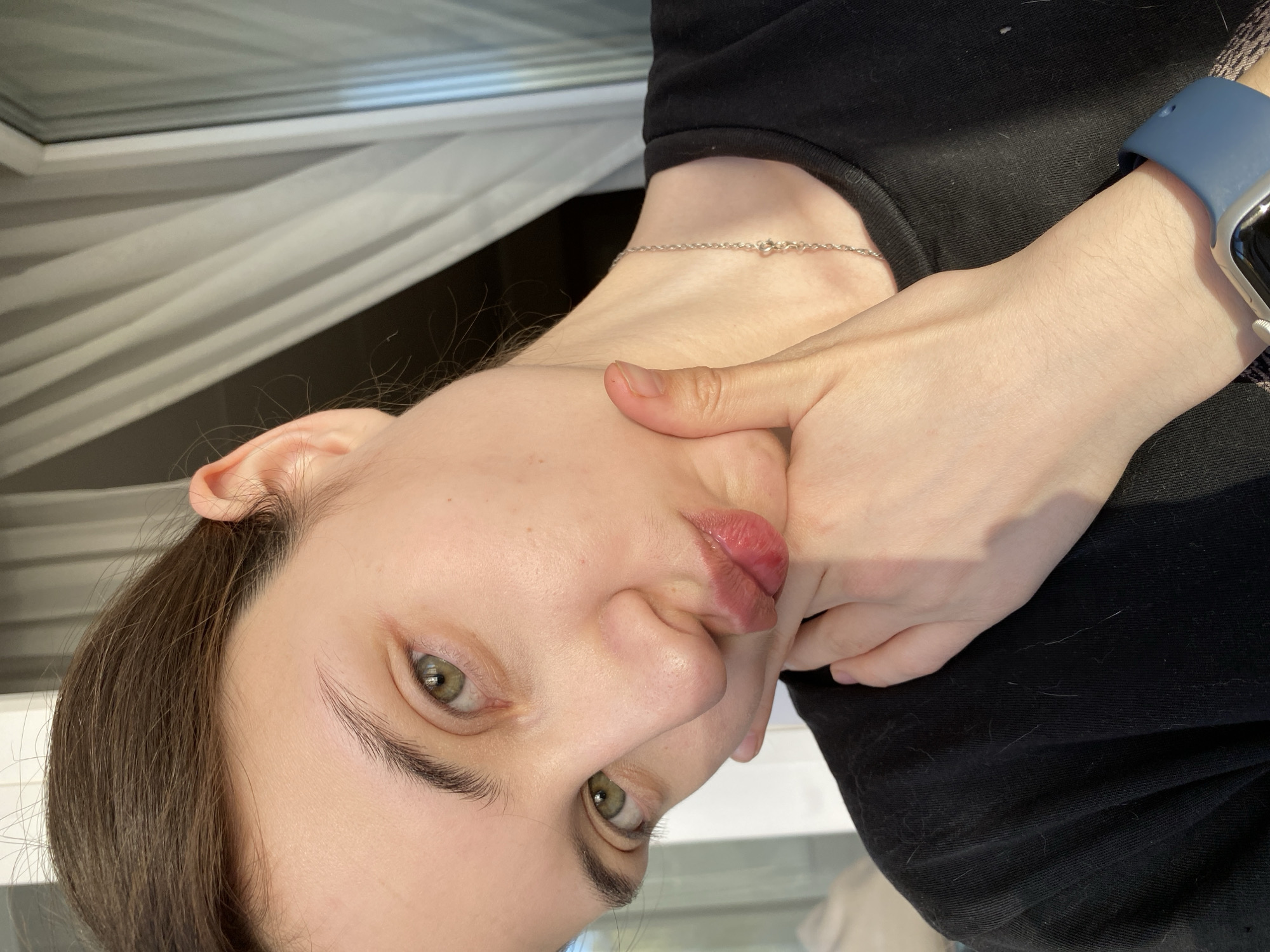 The image shows a person with fair skin, green eyes, and brown hair, holding their cheek with their hand in an upward angle selfie. They are wearing a black shirt and a silver necklace. A blue smartwatch is visible on their wrist. In the background, there is a window with white curtains. The location cannot be determined to be Votkinsk, Russia, or any other specific place.