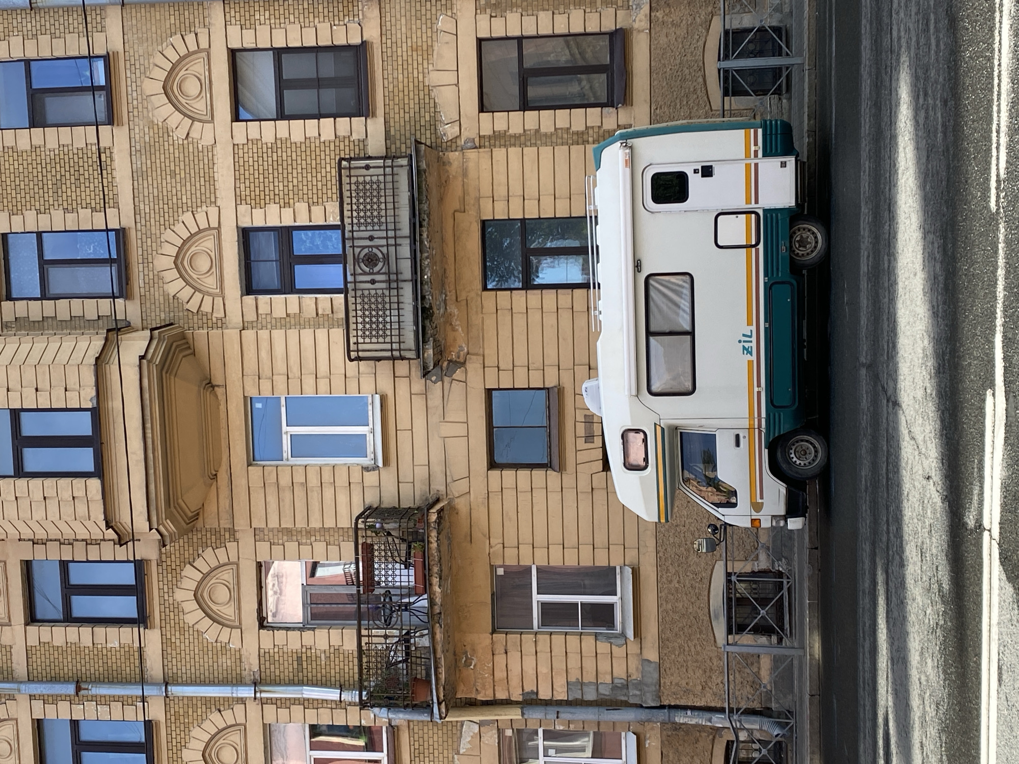 An overhead shot of a street in Saint Petersburg, Russia, shows a building with multiple windows, some with balconies, constructed of beige brick. Parked next to the building is a white and green recreational vehicle with the word "ZIL" printed on its side. The street is asphalt, with visible lane markings.