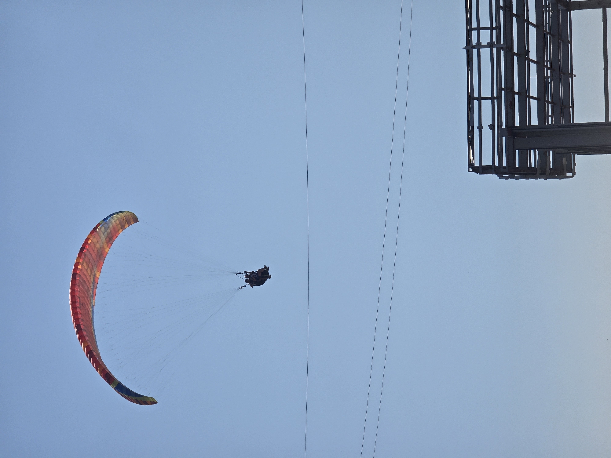 A tandem paraglider, with two people seated in a harness suspended beneath a colorful wing, soars against a clear blue sky. The paraglider is positioned in the upper left quadrant of the image. In the lower right quadrant, a portion of an unfinished, industrial-looking structure, possibly a billboard frame or scaffolding, is visible. Thin, dark lines, likely power lines or cables, traverse the sky horizontally across the frame. The scene is devoid of any specific landmarks that would pinpoint a precise location within Brajići, Montenegro.