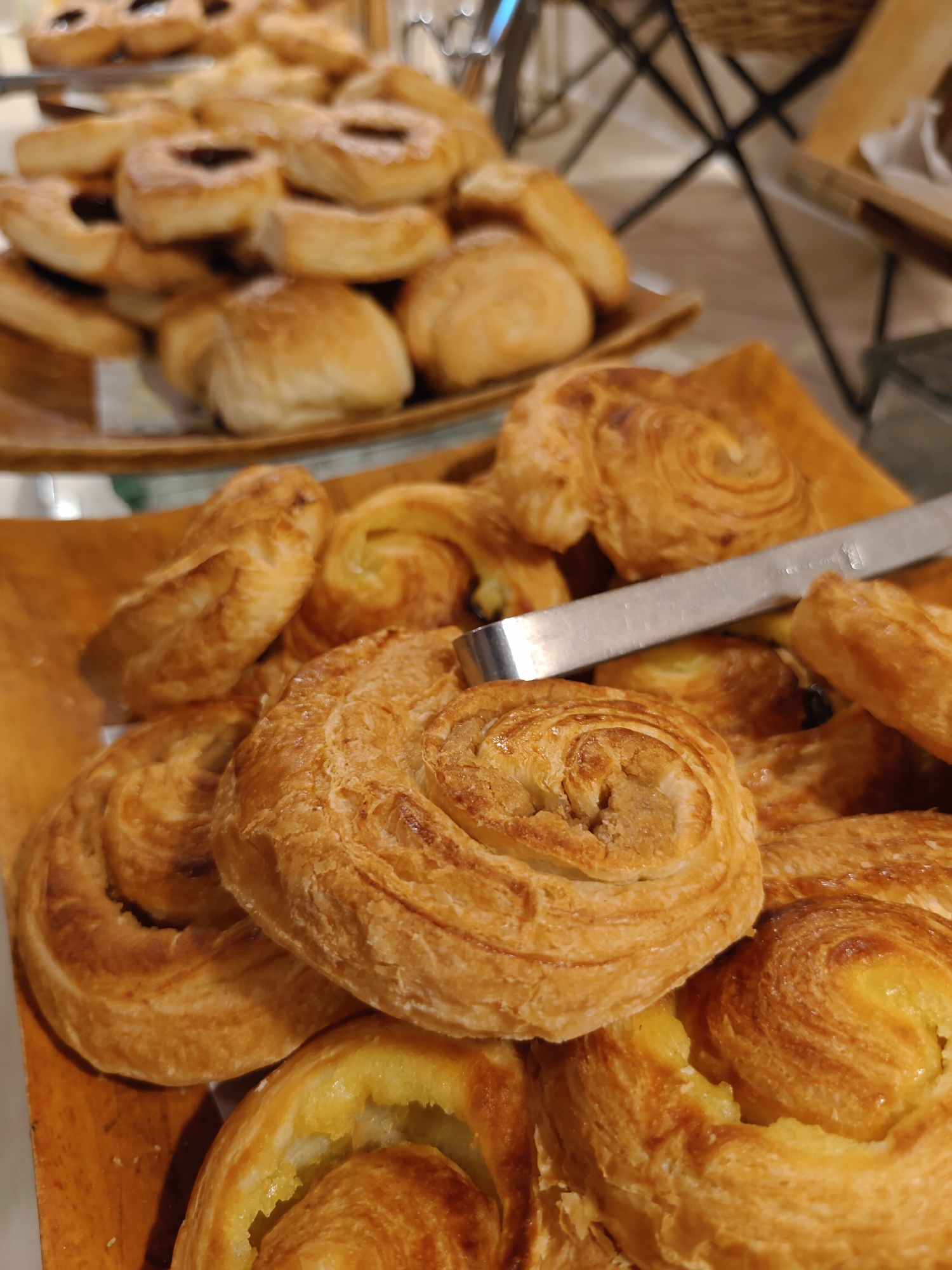 The image shows a close-up view of a selection of pastries, likely from a buffet or bakery, placed on a wooden tray. The pastries are golden brown and appear to be flaky and sweet. In the foreground, several swirled danishes with visible filling are prominent. Behind these, other pastries, including some with dark filling and round, bread-like rolls, are partially visible. A metal serving tong rests on the pastries. The background is blurred but suggests an indoor setting with some decorative elements. The location is identified as Pattaya City, Thailand.