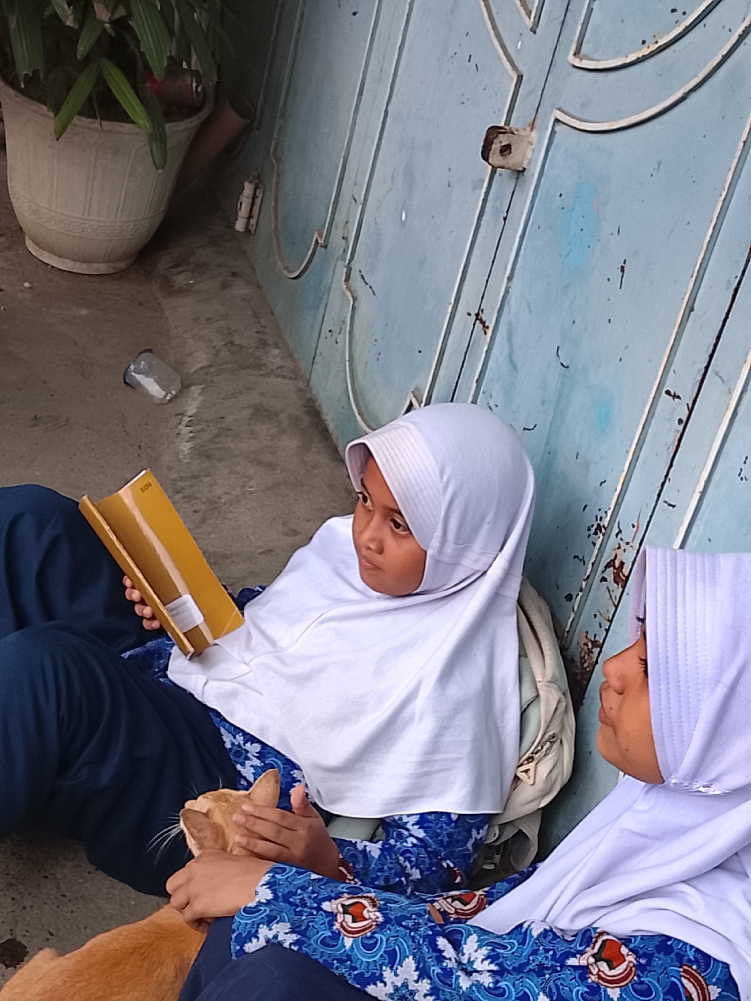 Two young girls, likely students, are seated on the ground outdoors, leaning against a worn, light blue metal door or wall panel. Both girls wear white hijabs, dark blue trousers, and blue patterned school uniform tops.
One girl, positioned on the left, holds an open yellow book or folder, possibly a notebook, which has the word "SIDU" visible on its spine. She looks off to her right with a calm, slightly pensive expression. A light-colored backpack is visible behind her.
The other girl, on the right, is partly visible, looking towards the first girl. Her hands are engaged in petting a light orange (ginger) cat that rests on her lap and the first girl's lap. The cat appears relaxed under their touch.
In the background, to the far left, a large, light-colored decorative pot with a green plant is visible. On the concrete ground near the girls, a small, clear plastic bottle or cup lies on its side. The scene suggests a daytime setting with natural light, implying pleasant weather. The overall atmosphere is one of casual rest or a break, possibly after school. The attire and brand name "SIDU" are consistent with the provided location context of Lhokseumawe, Indonesia.