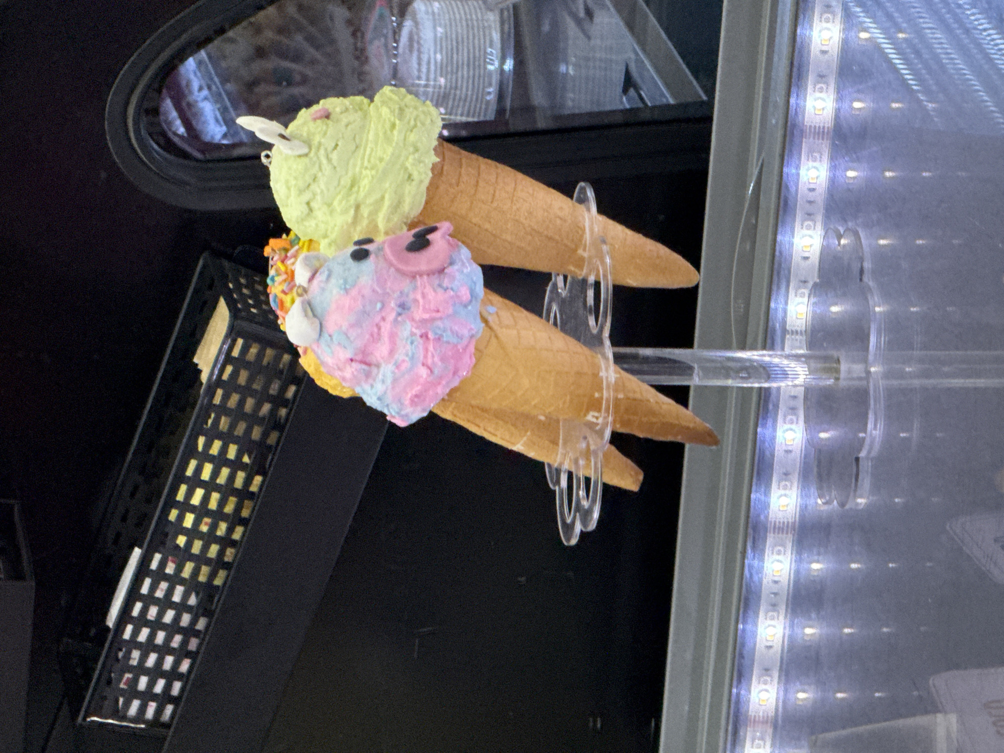 Two ice cream cones are displayed on a transparent stand inside what appears to be a refrigerated display case. The cone on top has yellow ice cream with a white candy decoration resembling wings. The lower cone features multi-colored ice cream, with a small pig-shaped candy on top and colorful sprinkles on the side. A black, perforated basket-like container is partially visible on the left side of the image. A strip of LED lights runs vertically along the right side of the display case. The scene likely takes place in a shop selling ice cream, but the precise location in San Bernardino Tlaxcalancingo, Mexico is impossible to determine from the image.