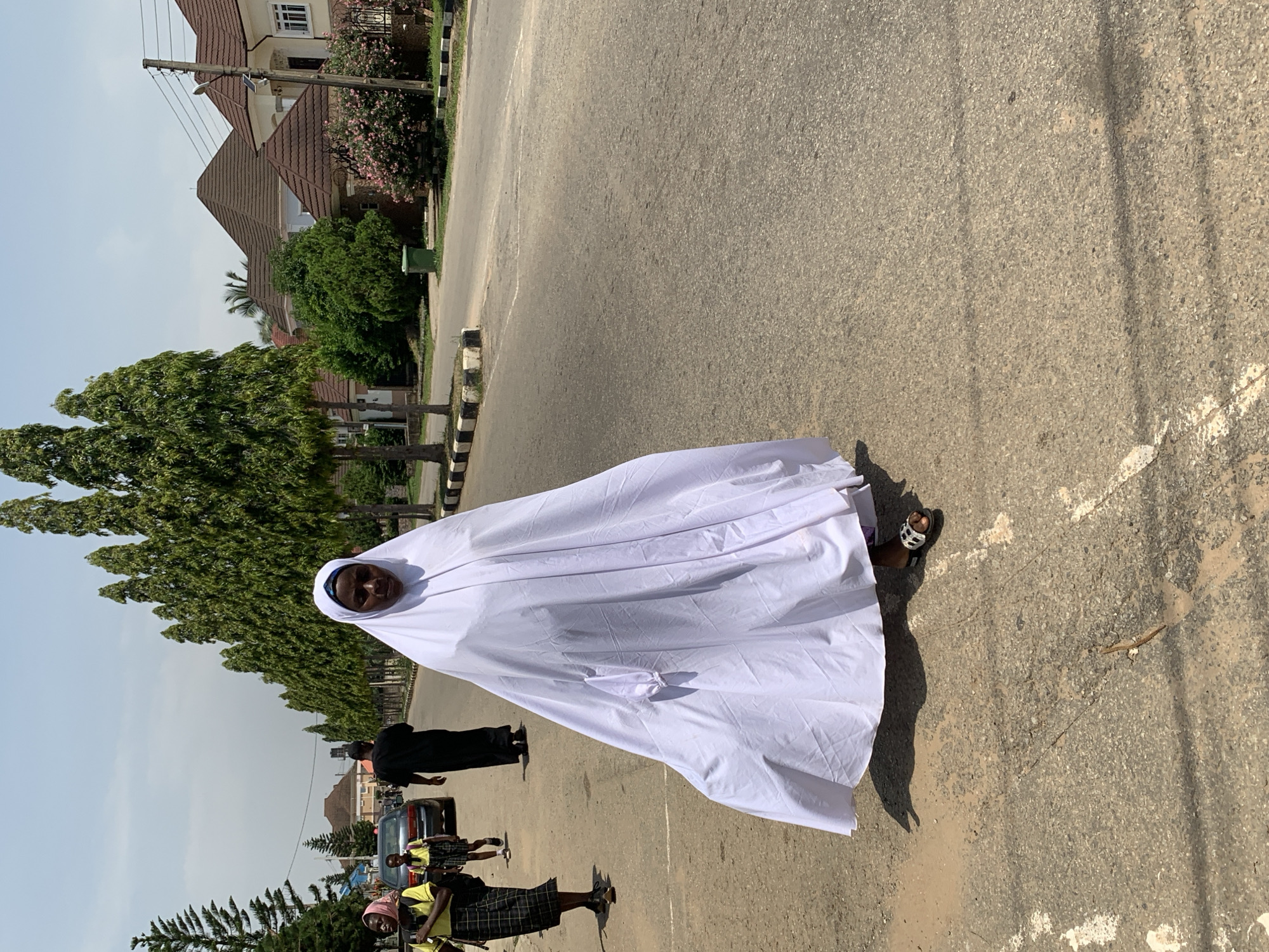 An overhead shot captures a woman walking on a paved street in Abuja, Nigeria. She is covered from head to toe in a white garment, with only her face visible. The woman wears patterned sandals on her feet. In the background, several other individuals are walking, including two young girls in school uniforms. A dark-colored car is also visible on the street. The street is lined with mature trees and houses with brown tiled roofs. The sky is clear and bright, indicating a sunny day.