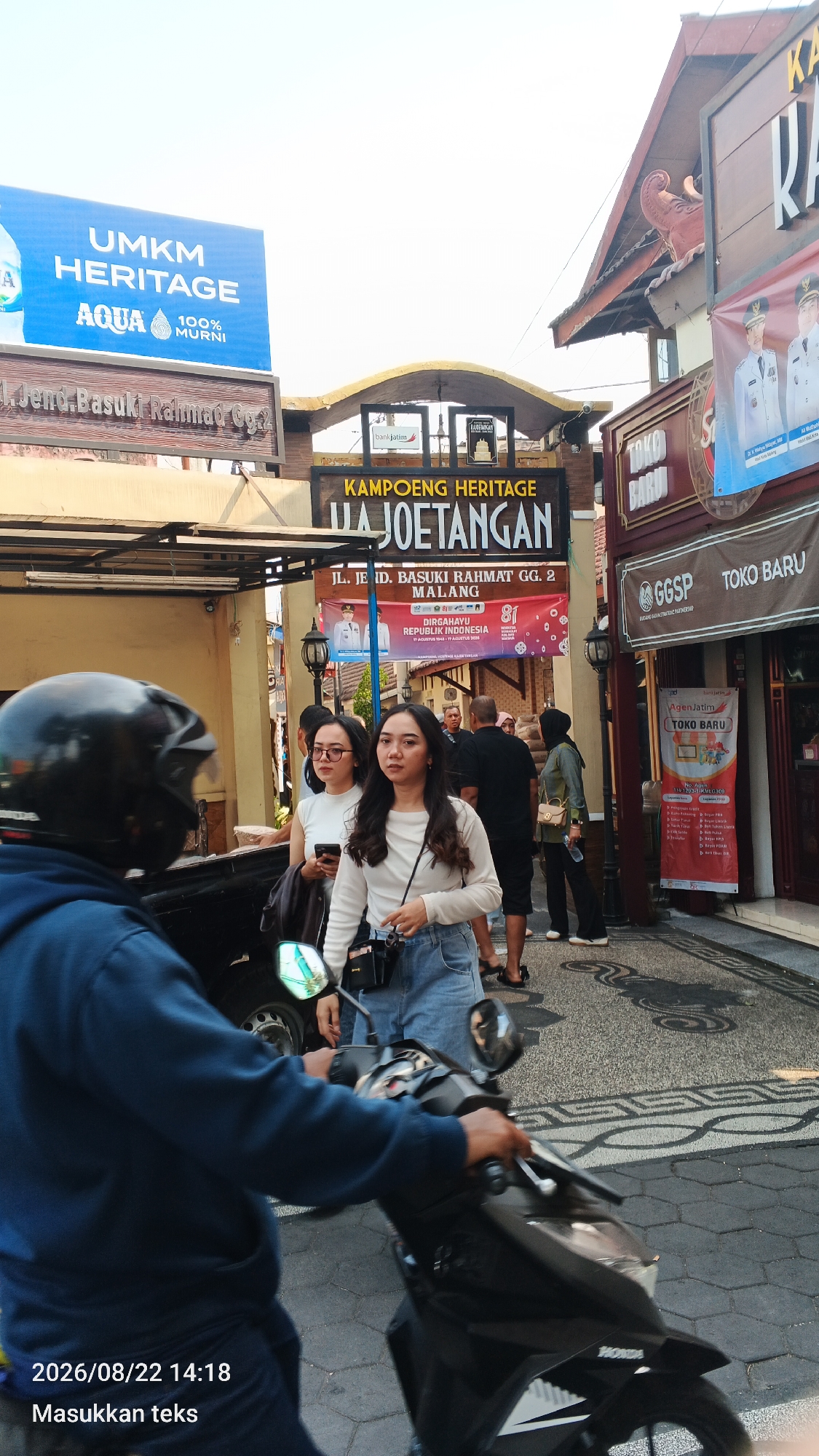 The image captures a street scene in Malang, Indonesia, likely in the heritage district known for its historical architecture and vibrant atmosphere. In the foreground, a person wearing a dark blue hoodie rides a black scooter, their body partially obscured by the vehicle. The scooter has a Honda logo visible on its front.
Behind the scooter, two young women stand on the sidewalk. The woman on the left wears glasses and a white top, holding a smartphone. The woman on the right wears a white long-sleeved shirt and denim shorts, with a camera hanging from her neck.
Above them, several signs are displayed. A prominent blue sign reads "UMKM HERITAGE AQUA 100% MURNI". Below this, another sign indicates "Jl. Jend. Basuki Rahmat Gg. 2 MALANG". A larger sign with "KAMPOENG HERITAGE" and "UA JOETANGAN" is also visible. Further to the right, signs for "GGSP TOKO BARU" and "AgenJatim TOKO BARU" are displayed. A banner featuring two men in uniform, possibly government officials, and text related to Indonesian Independence Day is also present.
The street is paved with cobblestones and decorative patterns. The buildings on either side of the street have traditional architectural elements, including wooden eaves and ornate details. The sky is bright and clear, suggesting a daytime setting. The overall impression is of a lively urban environment with a blend of commercial activity and cultural heritage.
