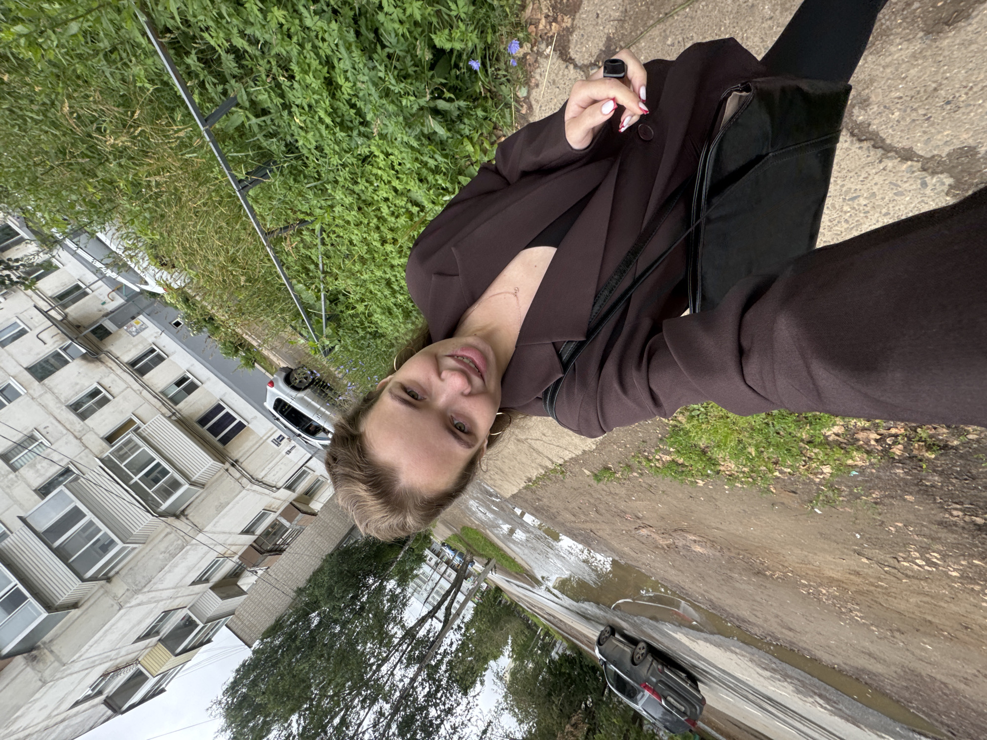 A young Caucasian female is taking a selfie outdoors, standing on a sidewalk next to a muddy road with puddles. She is wearing a dark brown blazer, a black V-neck top, and black pants. A black purse hangs from her left shoulder. Her hair is light brown and pulled back. She is wearing hoop earrings and a delicate necklace. Her right hand is visible, holding a black ring. In the background, there is a multi-story apartment building, trees, and a metal railing bordering overgrown green vegetation. A silver car is parked near the building, and a dark gray car is visible in the road. The scene appears to be in a residential area of Chaikovsky, Russia, likely on a cloudy or overcast day.