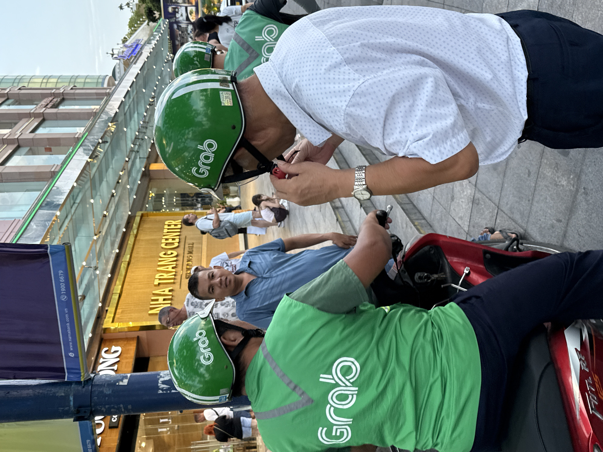 The image depicts a street scene in Nha Trang, Vietnam, specifically in front of the Nha Trang Center shopping mall. Two men wearing green Grab helmets and vests are the central figures. The man on the left is seated on a red motorcycle, facing away from the camera, with the Grab logo clearly visible on his vest and helmet. The man on the right, wearing a white patterned shirt and dark pants, is standing and appears to be adjusting something red near his mouth, possibly a mask or a device. His watch is also visible. Other people, some wearing Grab gear, are in the background, as well as shoppers and pedestrians. The architecture of the Nha Trang Center, with its modern facade and glass awnings, is prominent. The overall atmosphere suggests a busy urban environment, likely during the day.