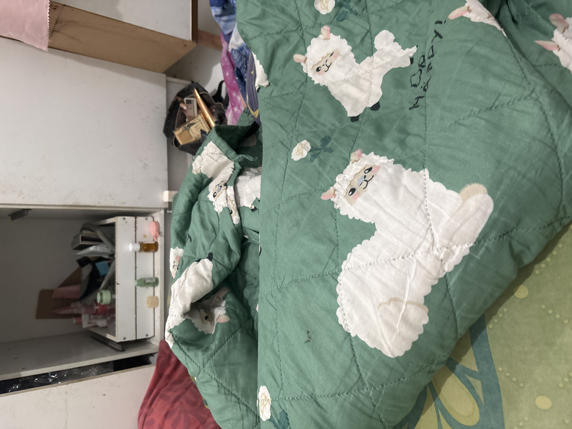 The image depicts a close-up view of a green quilt with a pattern of white cartoon llamas. The quilt is draped over a bed. In the background, a shelf in a cabinet is visible, containing various personal care items and a black bag with makeup. The setting appears to be an indoor residential space, possibly a bedroom. There are no people visible in the image. The overall scene suggests a personal space with everyday objects. The prompt specifies the location as North Jakarta, Indonesia, but this information cannot be visually confirmed from the image itself.