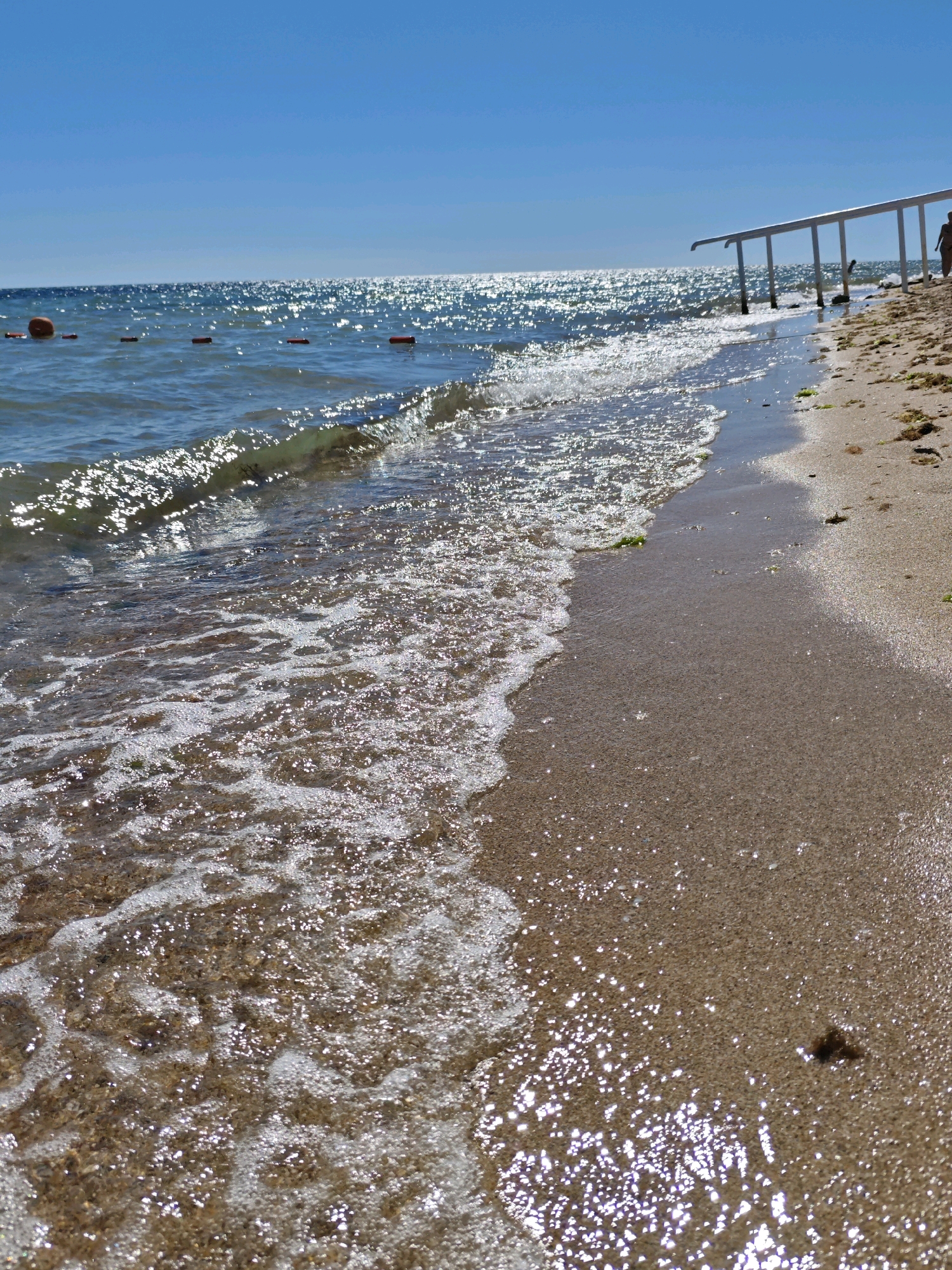 A low-angle, wide shot captures a sunny day at the beach in Yevpatoriya, Ukraine. The scene is dominated by the azure sky and the shimmering, sun-drenched sea. Gentle waves lap the sandy shore, leaving a foamy trail. In the distance, a white railing structure stretches along the beach, and a solitary figure can be seen walking near it. Orange buoys are also visible in the water, marking a swimming area. The sand is a light brown, with scattered debris from seaweed. The overall impression is one of a peaceful, sunlit day at the seaside.