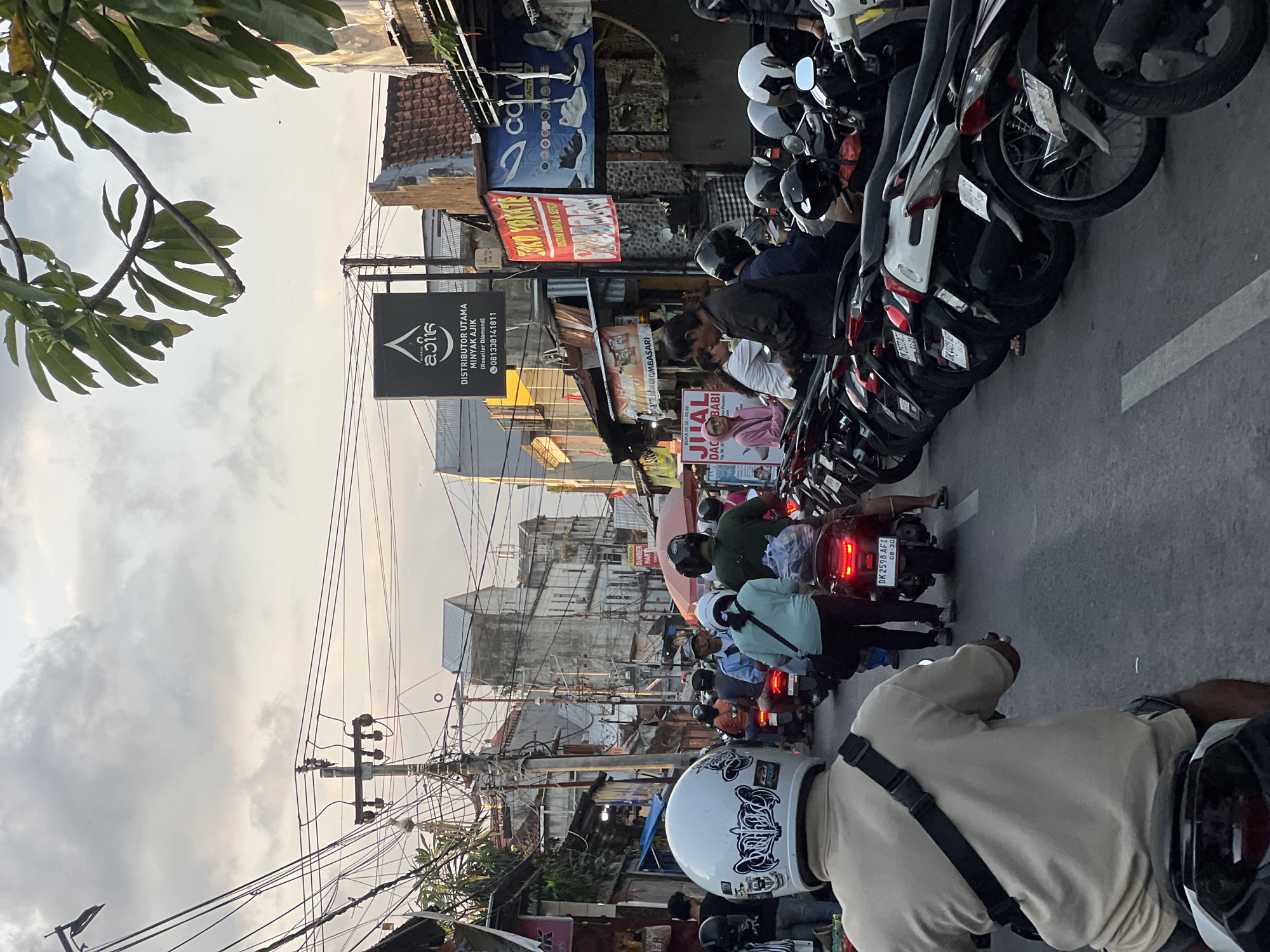 The image captures a bustling street scene in Denpasar, Indonesia, likely during a time of heavy traffic. The perspective is from behind a person on a motorbike, looking forward. In the foreground, the rider is wearing a white helmet with black and white graphics. The street is filled with a dense lineup of motorbikes parked along the right side.
On the left side of the street, a narrow lane is congested with motorbikes in motion. People are visible riding these motorbikes, some wearing helmets and others without. Shops and businesses line both sides of the street, displaying various signs and advertisements in Indonesian. One prominent sign features a logo and text that reads "AJIK MINYAK AJIK" with a phone number. Another sign advertises "TOKO YOKATE". Buildings appear to be multi-story structures, with some showing signs of wear and tear.
The environment is characteristic of a busy urban area in Indonesia, with a proliferation of electrical wires strung across poles overhead, creating a complex network. The sky is overcast with diffused light, suggesting it might be late afternoon or early evening. The overall impression is one of a vibrant and crowded marketplace or commercial district.