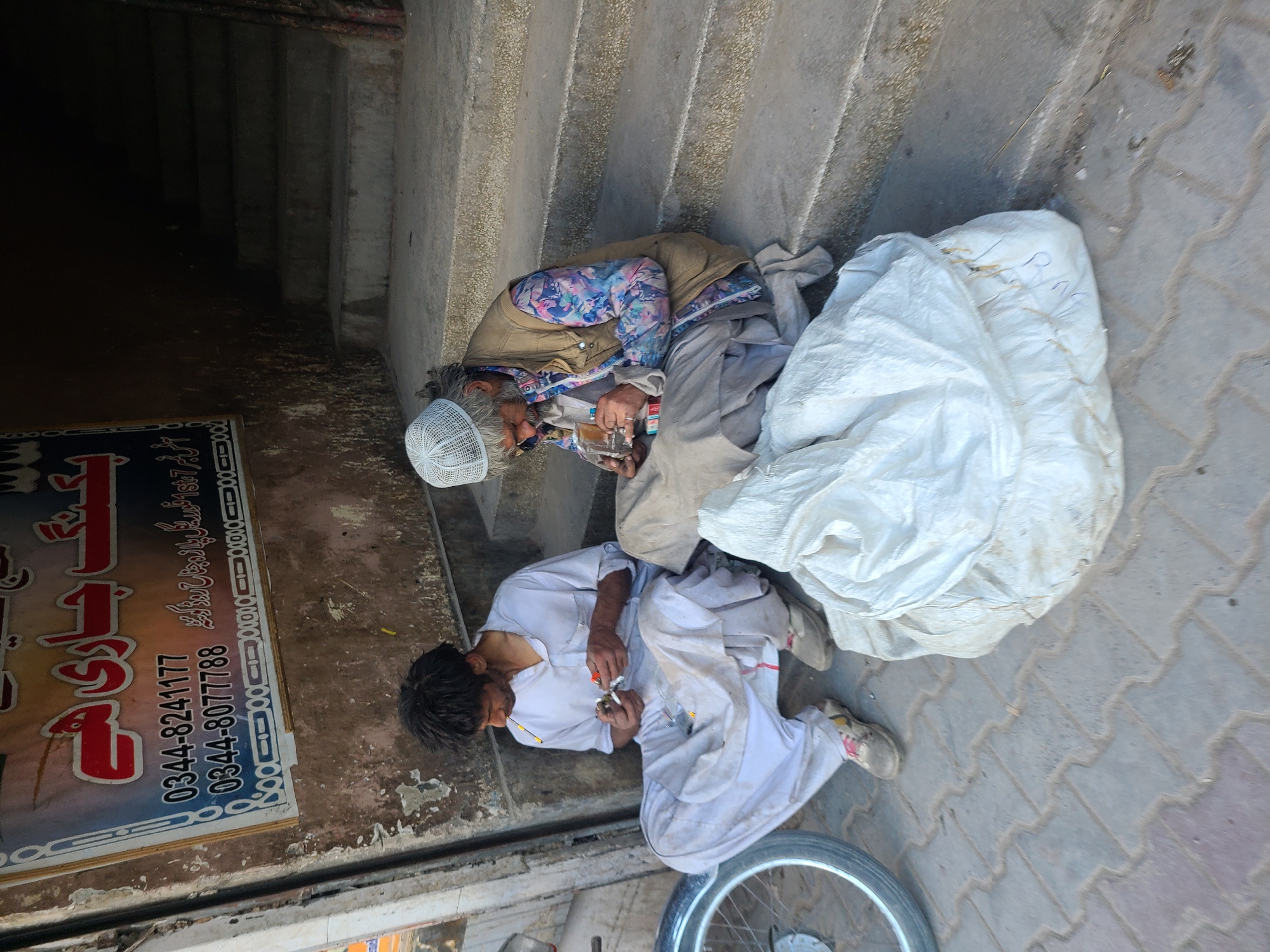 The image depicts two people next to each other on a street. One person appears to be sitting, wearing a beige jacket, light gray pants, and a white kufi. They are holding a clear container in their hands and seem to be using something to light it. The other person is lying on the ground, dressed in white traditional clothing. They seem to be lighting something with a small torch. Next to them is a bicycle wheel. Beside the seated person is a large white bag. There is a sign with Urdu script and phone numbers on the left side of the frame. The location appears to be in Quetta City Tehsil, Pakistan.