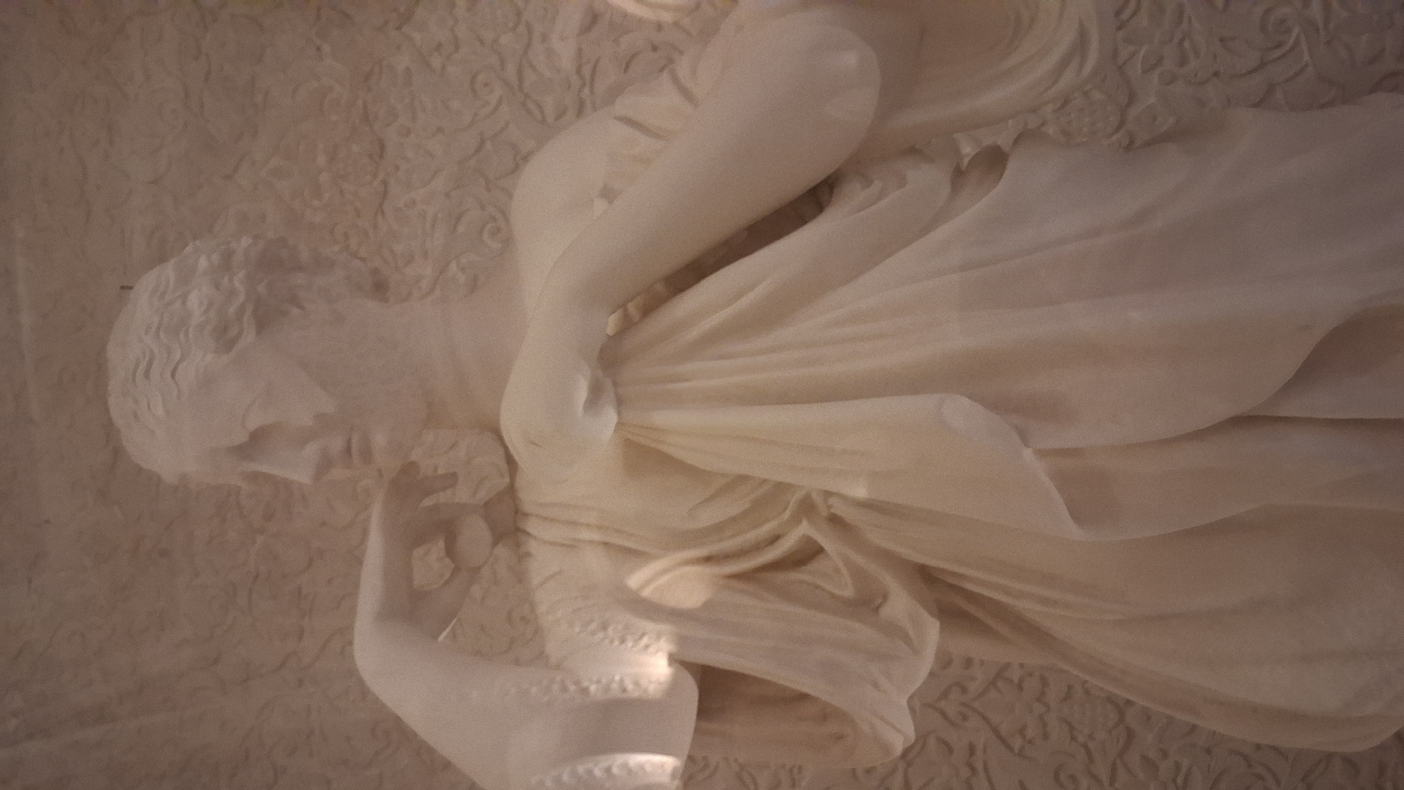 The image shows a close-up of a white marble statue of a woman, likely a classical figure, depicted from the waist up. The statue is positioned against a textured, light-colored background that appears to be a wall or panel with a decorative relief pattern. The statue is draped in flowing fabric that creates soft folds and highlights. One arm is bent, with the hand resting near the chin or cheek, and the other arm is extended outwards, also draped in fabric. The statue's head is turned slightly to the left, and her facial features are softly rendered. The lighting is somewhat dim, with a bright spot illuminating a portion of the fabric on the left side of the statue. The overall impression is one of grace and elegance. This statue could be found in the Quinta da Regaleira estate in Sintra, Portugal, which is known for its elaborate gardens and symbolic architecture.