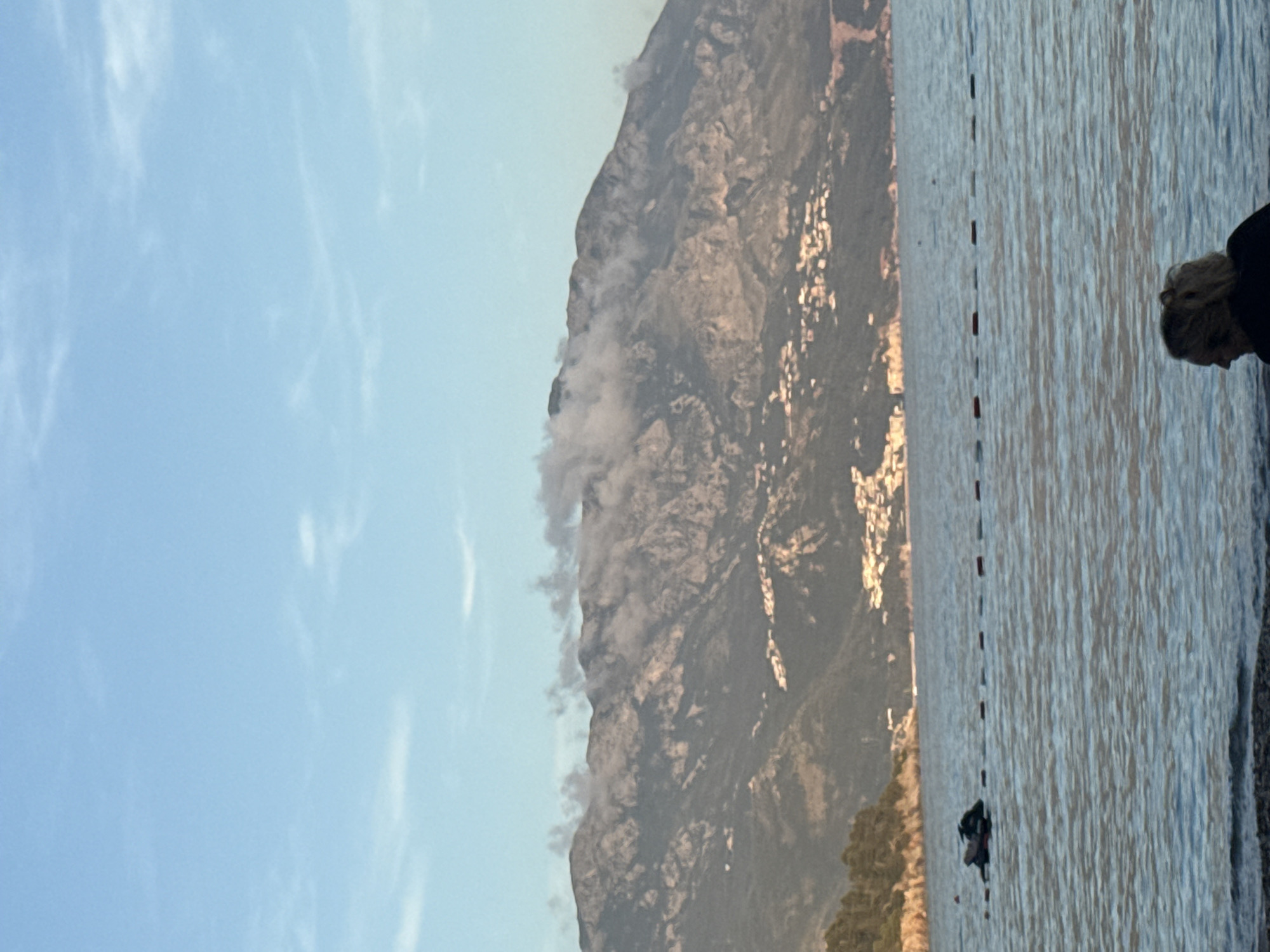 This image captures a serene coastal landscape in Budva, Montenegro, likely during the golden hour of either sunrise or sunset. A majestic, rugged mountain dominates the left side of the frame, its rocky slopes partially obscured by soft, wispy clouds or mist near its peaks. Sunlight, appearing golden, reflects brightly off various sections of the mountain, highlighting its contours.
Adjacent to the mountain, on the right, is a calm expanse of sea. The water's surface is gently rippled, reflecting the ambient light and creating shimmering patterns. A series of red buoys forms a distinct dotted line stretching across the water, indicating a boundary or swimming area.
In the foreground on the far right, the head and shoulders of a person are visible, seemingly observing the water or shoreline. Further out in the sea, near the line of buoys, a smaller, dark figure can be seen, possibly engaged in swimming or floating. The sky, which occupies the upper portion of the scene, is a clear, light blue, adorned with subtle, scattered white clouds. The overall mood conveyed is one of tranquility and natural beauty. There is no visible text in the image.