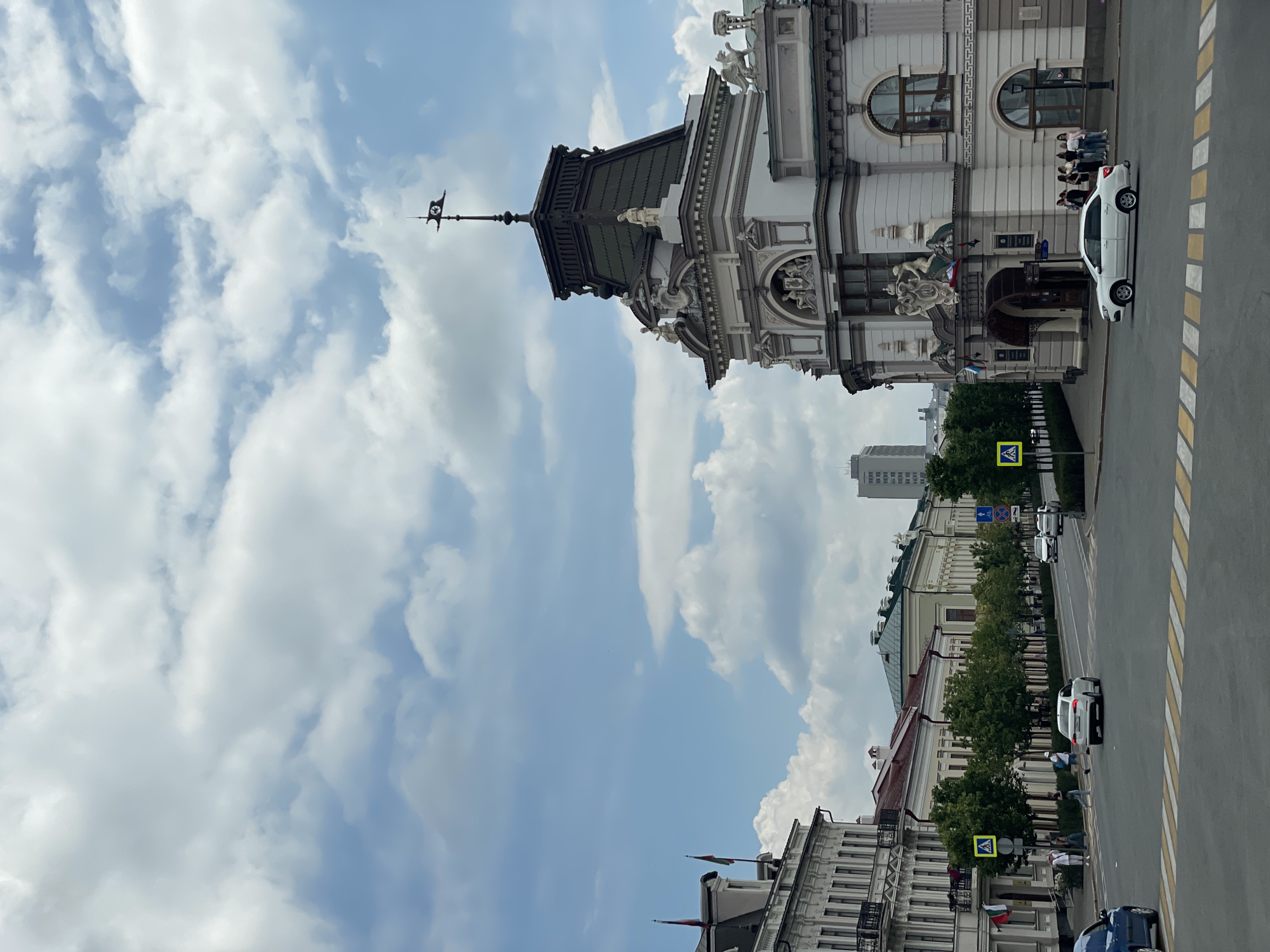 This image captures a view of the intersection of Bauman Street and Karl Marx Street in Kazan, Russia. The ornate architecture of the Kazan City Duma building is prominent on the right, with its distinctive tower topped by a weather vane. Across the street on the left, the elegant facades of historical buildings line Bauman Street. Several cars are visible on the road, including a white sedan crossing the pedestrian crossing in the foreground and a blue car on the far left. A few individuals are seen walking on the sidewalks and crossing the street. The sky is partly cloudy, with large, white cumulus clouds against a blue backdrop. Green trees line Bauman Street, adding to the urban landscape.