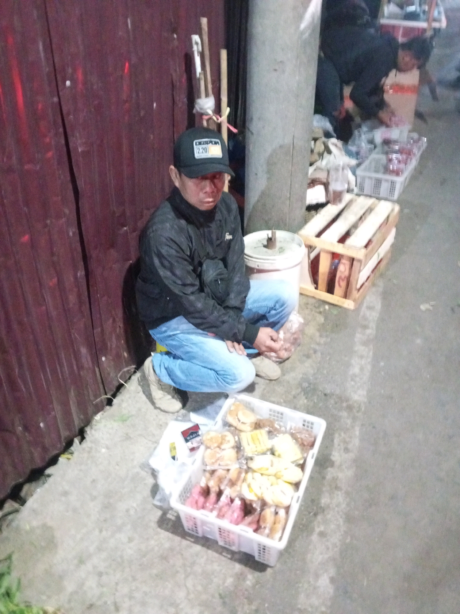 A man wearing a black baseball cap, black jacket, and blue jeans is crouching on a concrete sidewalk next to a dark red corrugated metal wall. He is looking down and appears to be handling something in his hands. In front of him, a white plastic basket contains various pastries, some of which are pink. To his right, a wooden pallet is visible, and further in the background, a child in a light-colored shirt is partially obscured. The scene appears to be outdoors, possibly at a street market or roadside stall in Gentan, Indonesia, as indicated by the context provided.