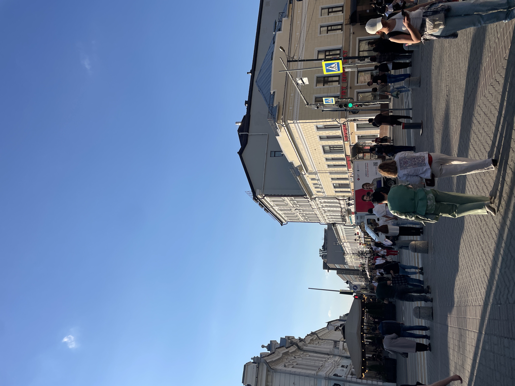 The image captures a street scene in Kazan, Russia, under a clear blue sky with a few scattered clouds. The perspective is slightly tilted. Buildings with ornate facades line the street. Pedestrians populate the sidewalk; some are walking, while others appear to be waiting at a crosswalk. The crosswalk has a pedestrian crossing sign and a green traffic light. Shadows cast by buildings and people are visible on the paved street. A restaurant or cafe with a tented awning is visible.