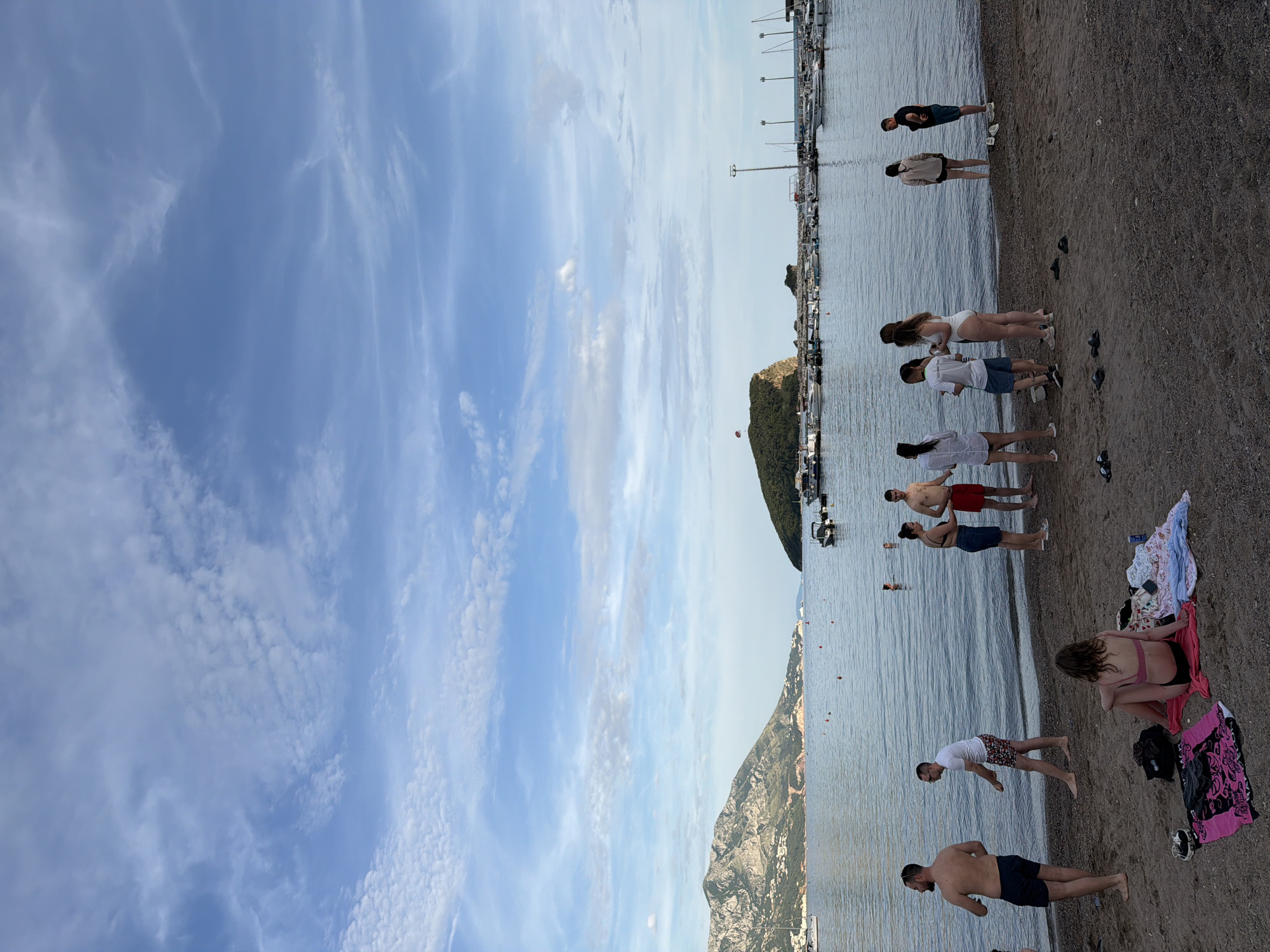 This image captures a wide view of a beach and bay in Budva, Montenegro, likely during the late afternoon or early evening. **Environment:** The sky is a clear light blue with scattered white and grey stratocumulus and cirrus clouds. The expansive bay features calm, dark blue-grey water. In the distance to the left, a large, green mountainous landmass with scattered white buildings extends along the horizon. Directly in the center-right of the bay, a prominent, verdant, rocky island (Sveti Nikola Island) is visible, topped with a small red flag. A red and white parasail is deployed from a boat near this island. To the right of the island, a marina is packed with numerous docked motorboats and sailboats, flanked by tall light poles. Several red buoys are distributed across the water. The immediate foreground consists of a dark, coarse sand and pebble beach. **People:** Approximately 15-20 individuals are present on the beach and at the water's edge. * On the far left, a shirtless man with a beard, wearing dark swim shorts, stands facing right. Adjacent to him, a man in a white t-shirt and patterned swim shorts leans towards the water. * Further right, a woman sits on a pink patterned towel on the sand, facing away from the camera, wearing a pink bikini top and dark bottoms. Clothes and a white towel are laid out beside her. Multiple pairs of sandals are scattered on the sand. * Near the water's edge, moving from left to right: * A group of three people stands: a woman in a dark swimsuit and dark shorts, a shirtless man in red swim shorts, and a woman in a white top and dark shorts. * Another cluster of three people includes a man in a white t-shirt and light patterned shorts, a woman in a white one-piece swimsuit, and a woman in a white top and dark shorts. * Towards the far right, two individuals stand at the water's edge: a woman in a light top and dark shorts, and a man in a black t-shirt and light shorts. * One person's head is visible partially submerged in the water in the mid-right, and another person stands in the shallow water further right. **Objects:** The beach area shows towels, clothing, and multiple pairs of sandals. The bay contains various recreational boats, including motorboats and sailboats, and red marker buoys. The marina is equipped