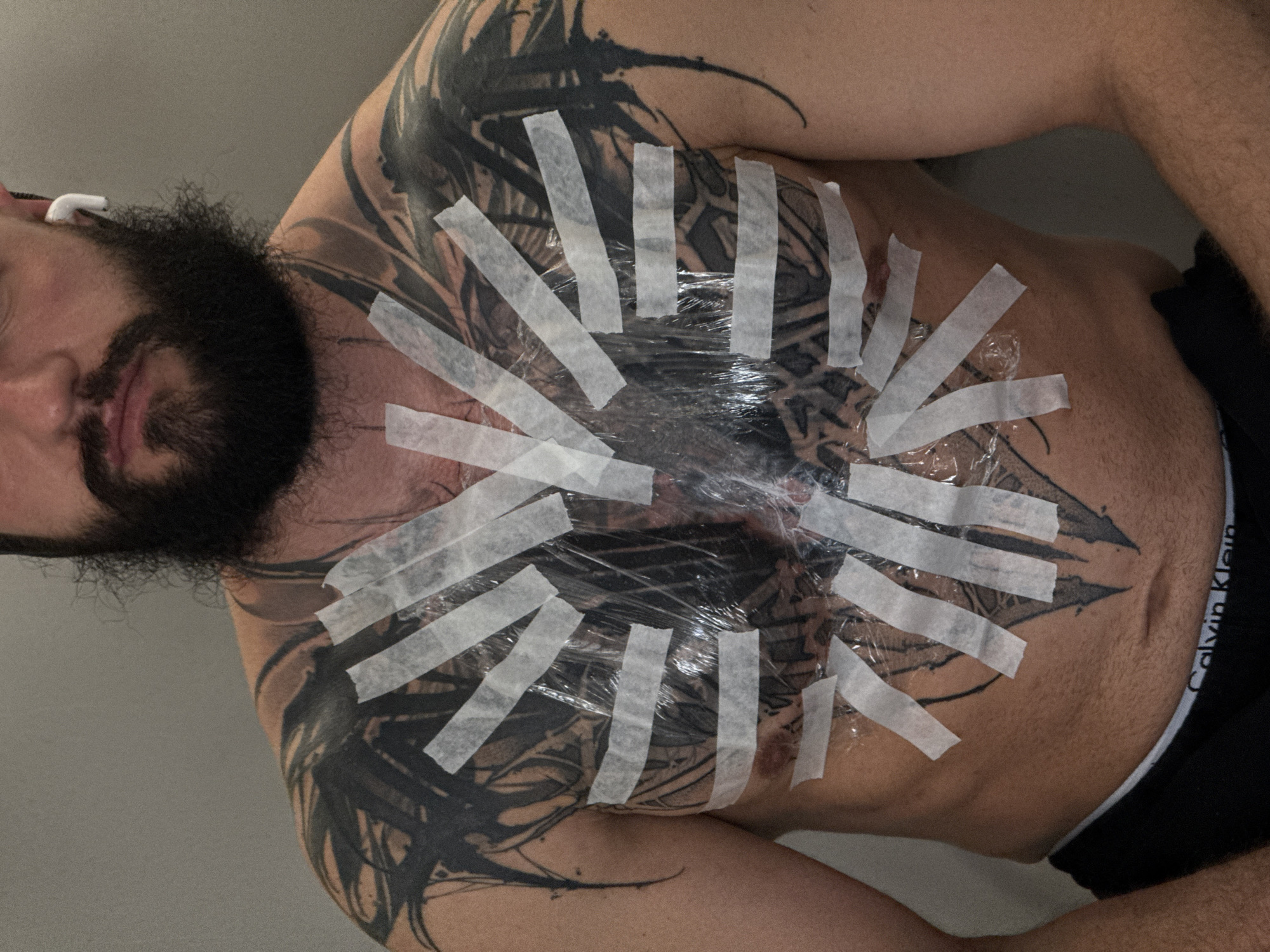 A medium shot, taken at an upward angle, depicts a man lying on his back. He has a full, dark beard and light skin tone. A white earbud is visible in his left ear. He is wearing black Calvin Klein underwear. His chest and shoulders are covered in an extensive black tattoo, which is covered with clear plastic wrap and secured with strips of white tape. The environment appears to be indoors, with a plain, light-colored wall visible in the background. The location is not discernible from the image.